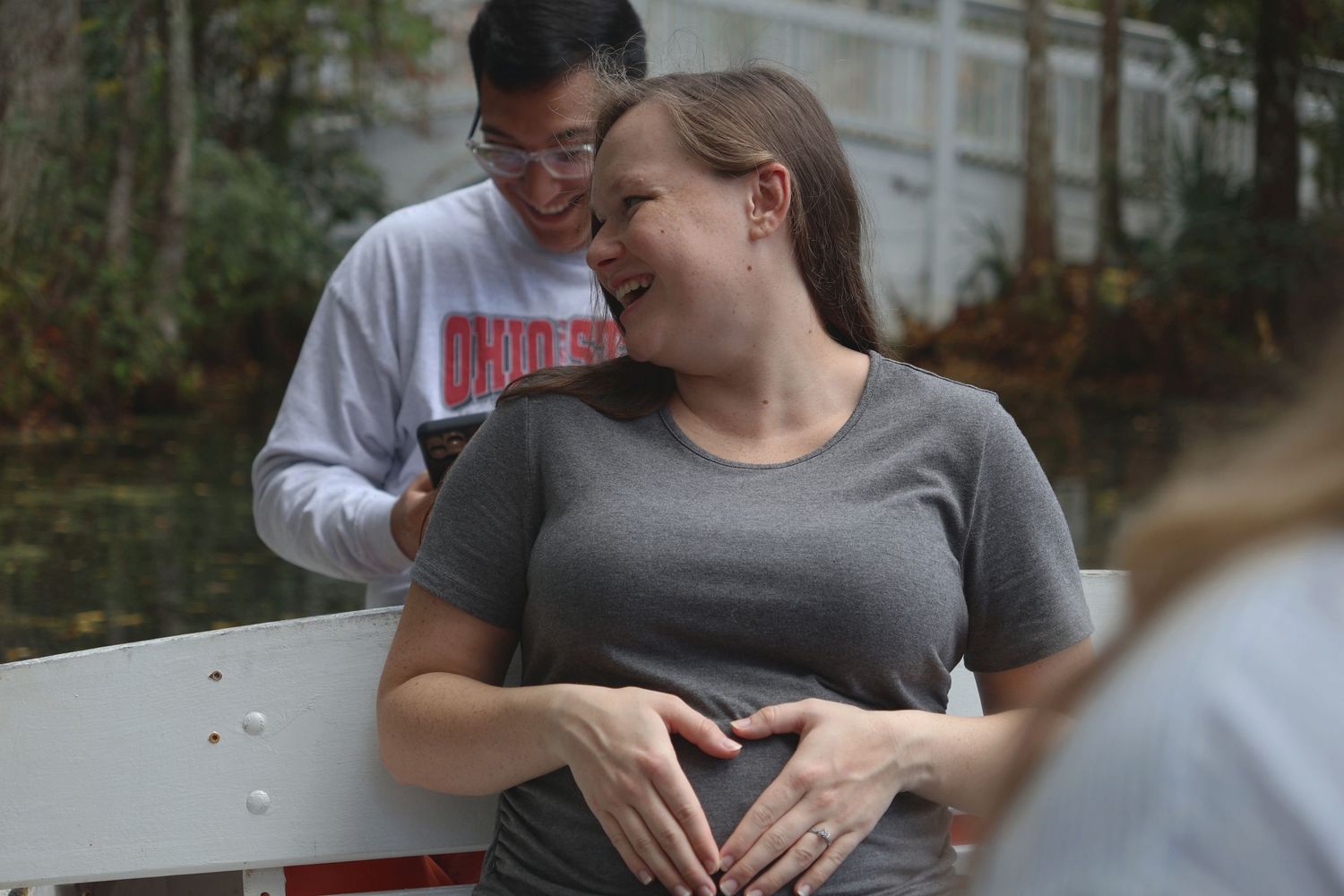 A laughing woman forming a heart with her hands over her stomach as a man smiles behind her in an outdoor setting.