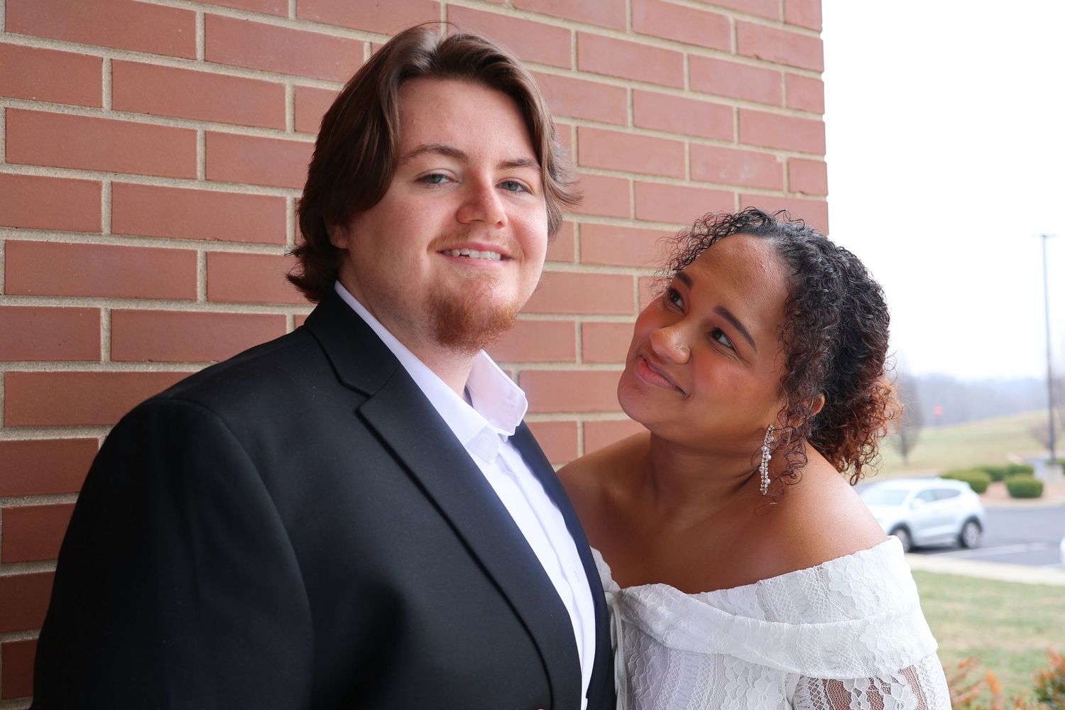 A groom in a black suit smiling at the camera while his bride in an off-the-shoulder dress looks up at him affectionately outdoors.
