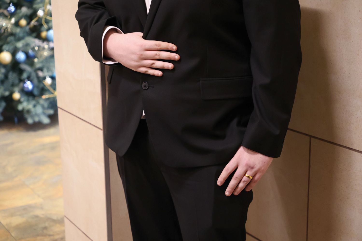 Close-up detail of a groom in a black suit resting one hand across his stomach and one hand in his pocket near a tiled wall.