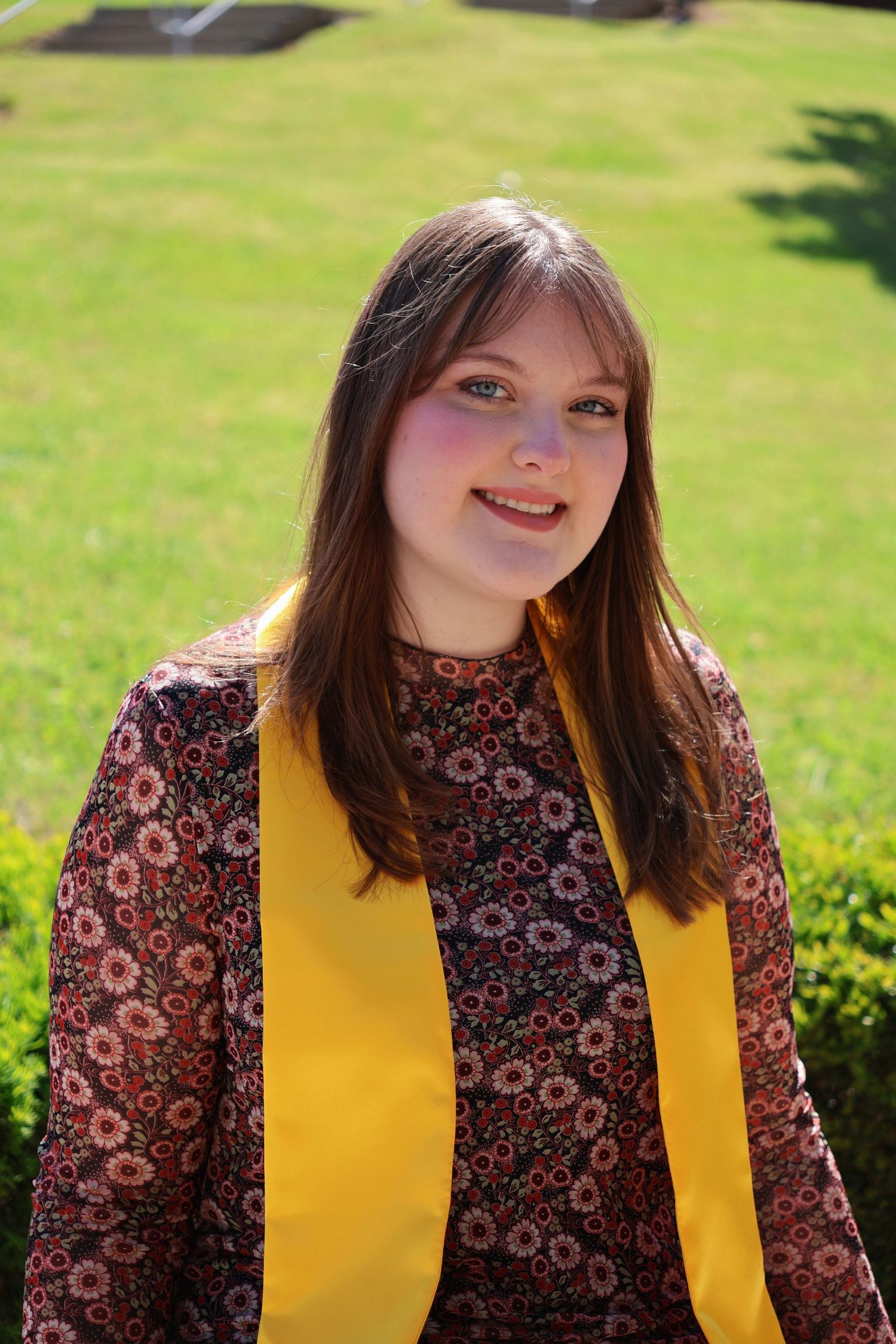A portrait of a smiling woman with bangs wearing a yellow graduation stole over a patterned dress outdoors on a sunny day.