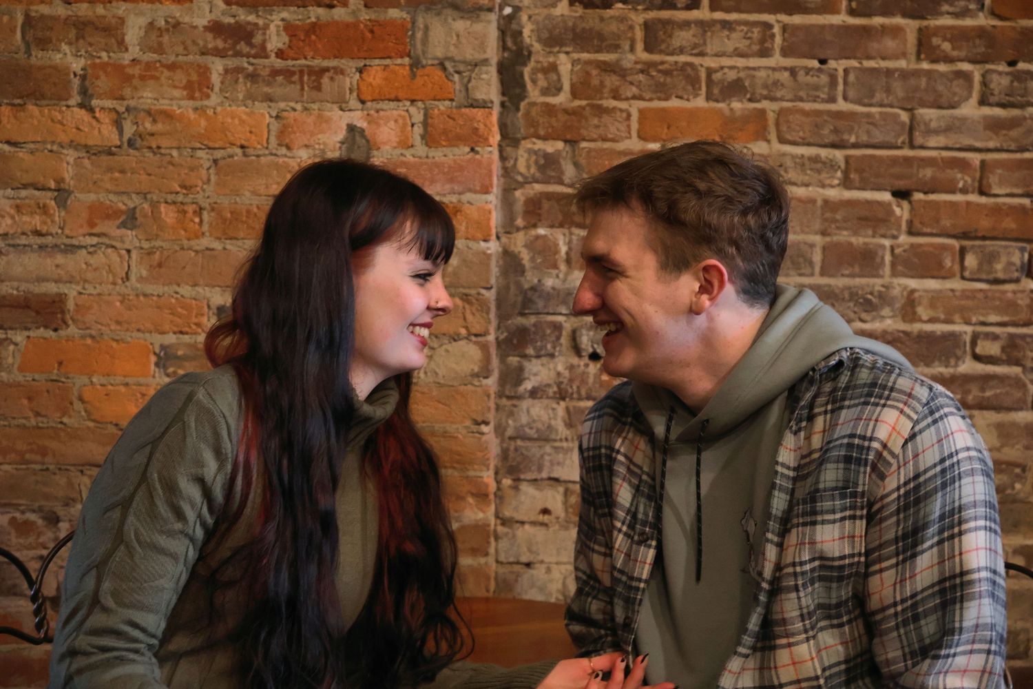 A smiling young couple sitting close together and laughing against an exposed red brick wall.