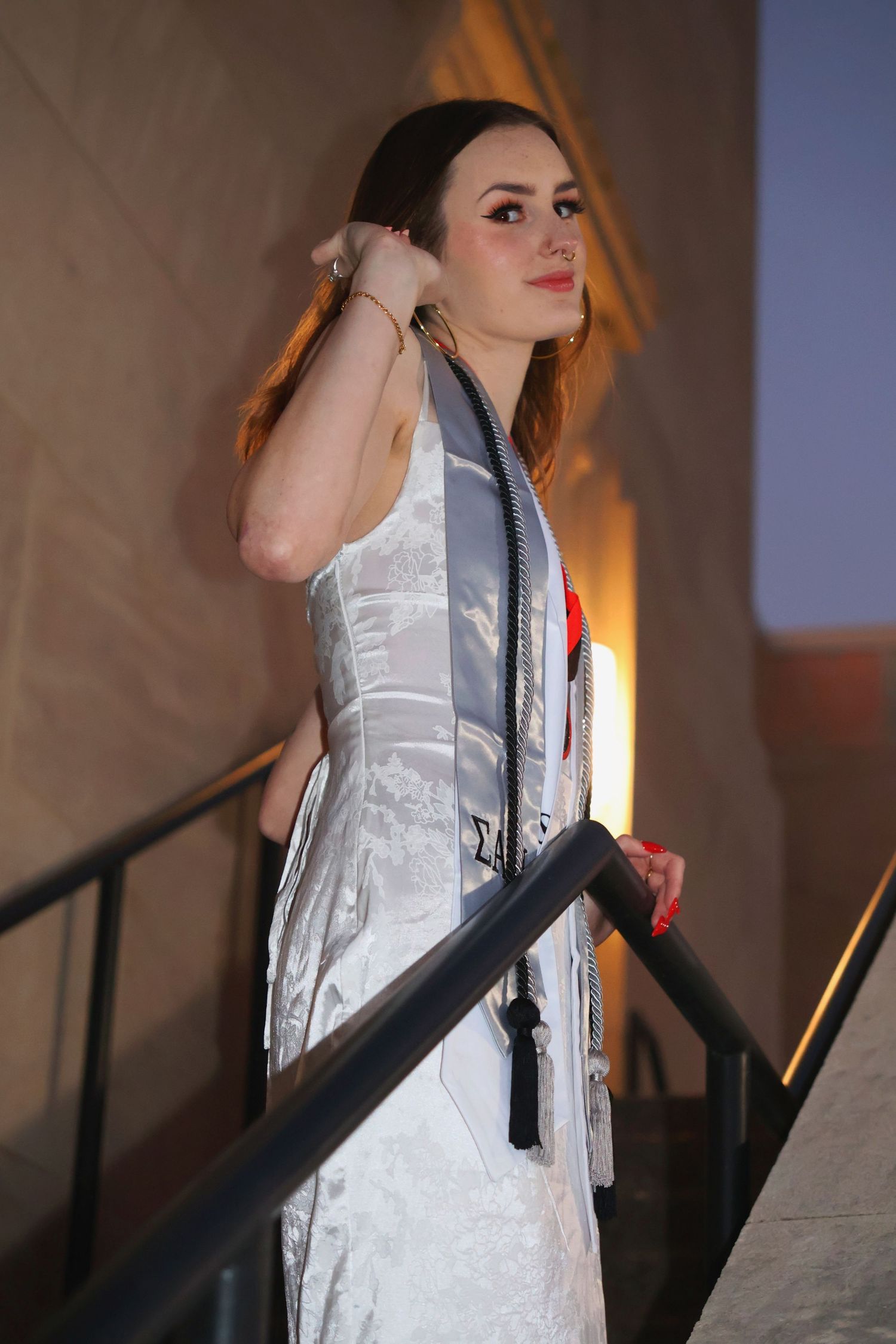 A woman in a white dress and graduation stole posing on an outdoor staircase with one hand near her hair and being backlit by a street lamp.