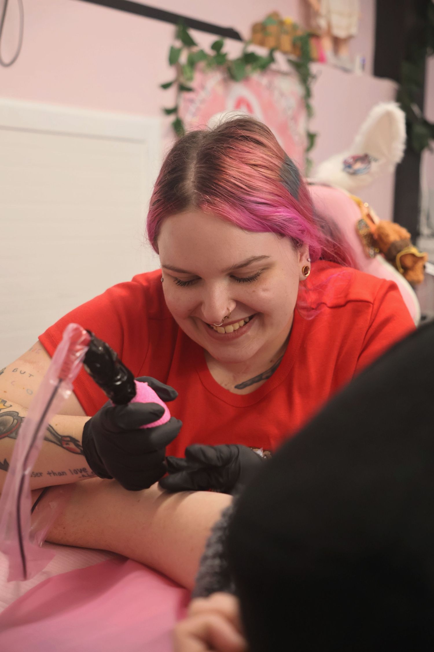 A smiling tattoo artist with pink hair and black gloves using a pink wrapped tattoo machine on a client's arm.
