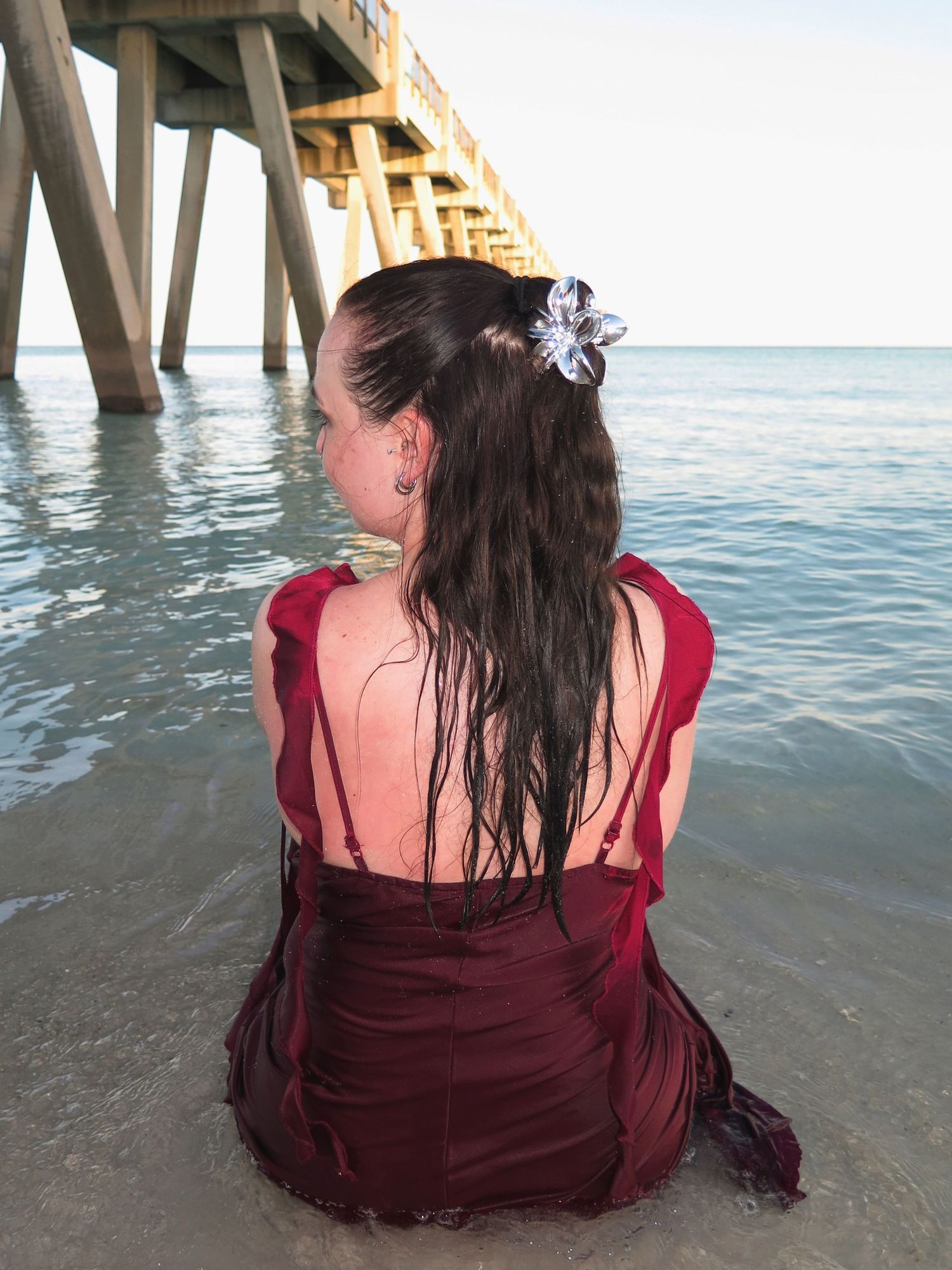 A woman with wet hair wearing a soaked maroon dress sitting on the sand facing toward a pier on the left.