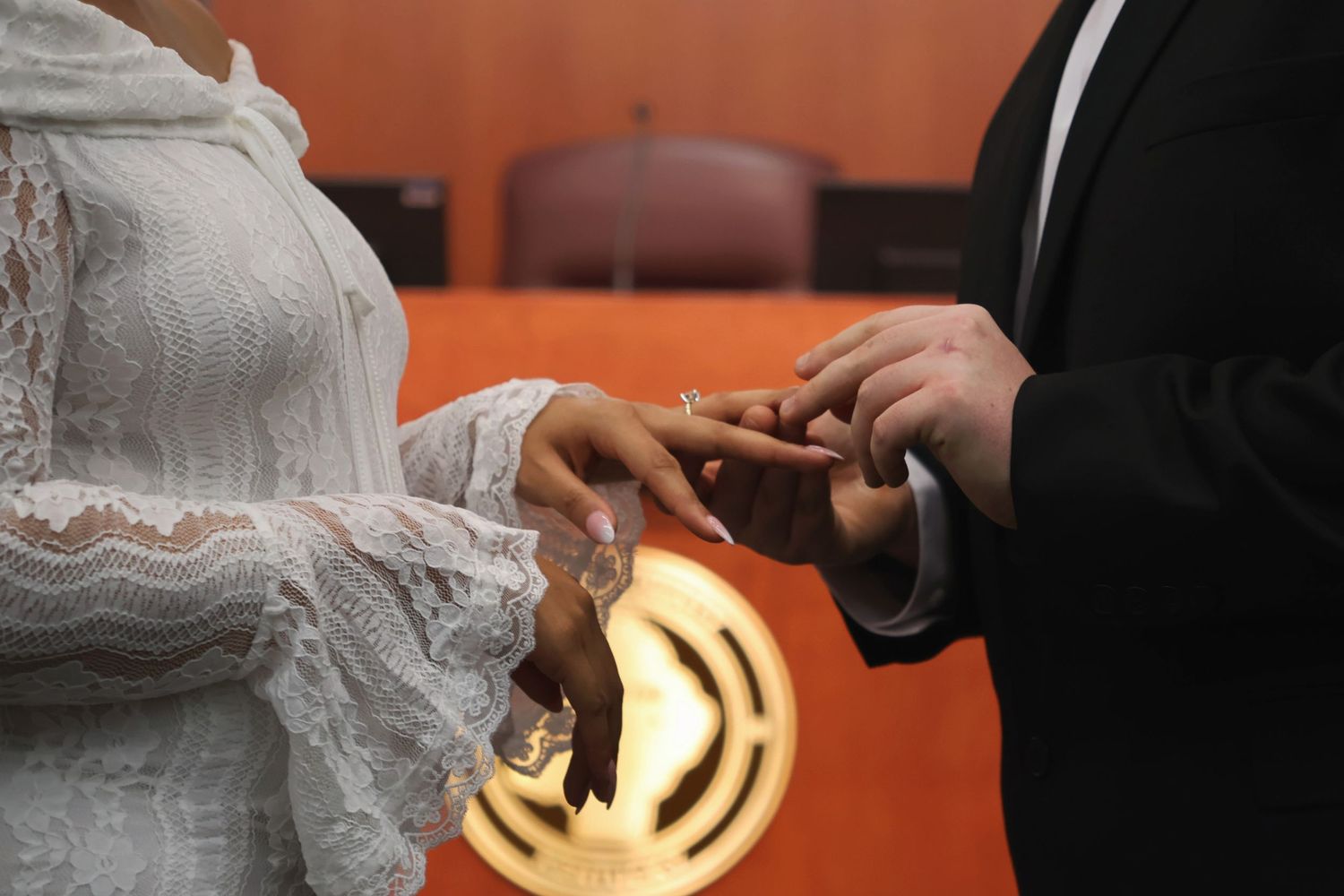 A close-up shot of a groom placing a wedding ring onto his bride's finger during a courtroom ceremony
