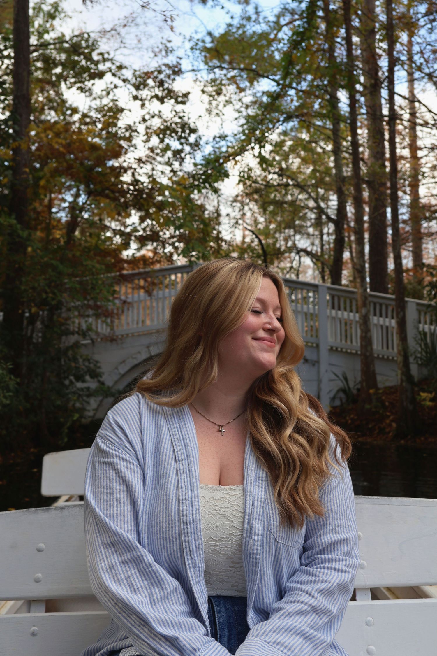 A smiling woman with blonde hair wearing a light blue unbuttoned shirt sitting outdoors with autumn trees in the background.