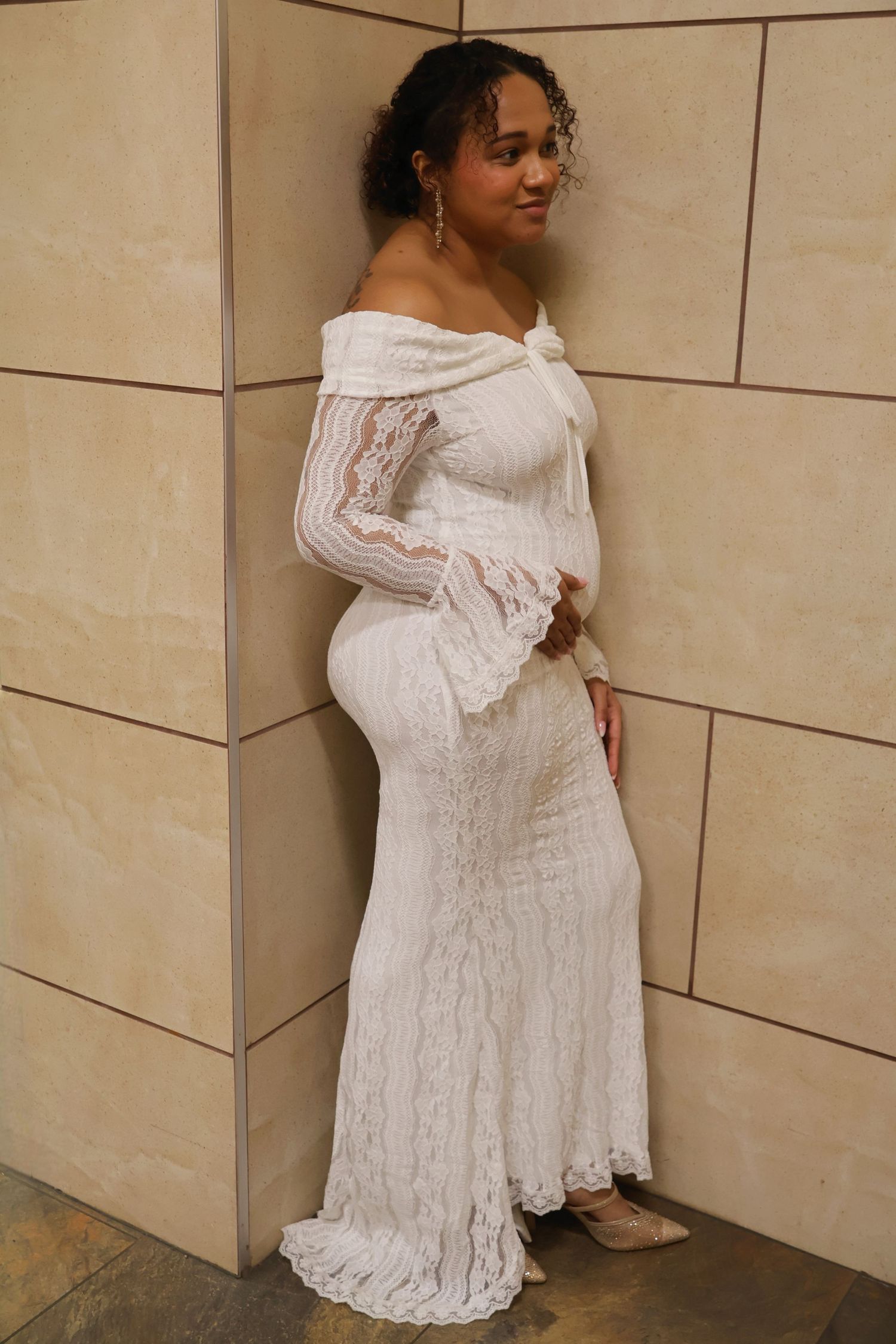 Full-length portrait of a bride in a white off-the-shoulder lace gown with long bell sleeves standing against a tiled wall.