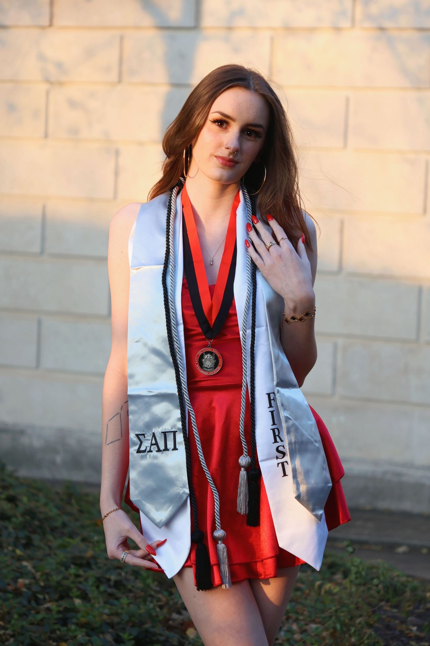 A woman graduate wearing a stole leaning against a wooden bridge railing with autumn trees in the background.