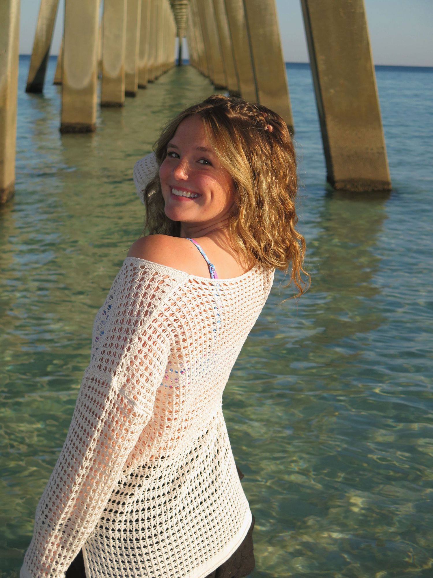 A smiling woman in a open-knit white sweater looking back over her shoulder while standing in shallow ocean water under a pier.