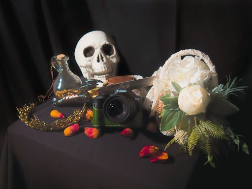 A dark still life arrangement with a human skull, a camera, a glass bottle, a crown, scattered flower petals, & a basket of white florals.