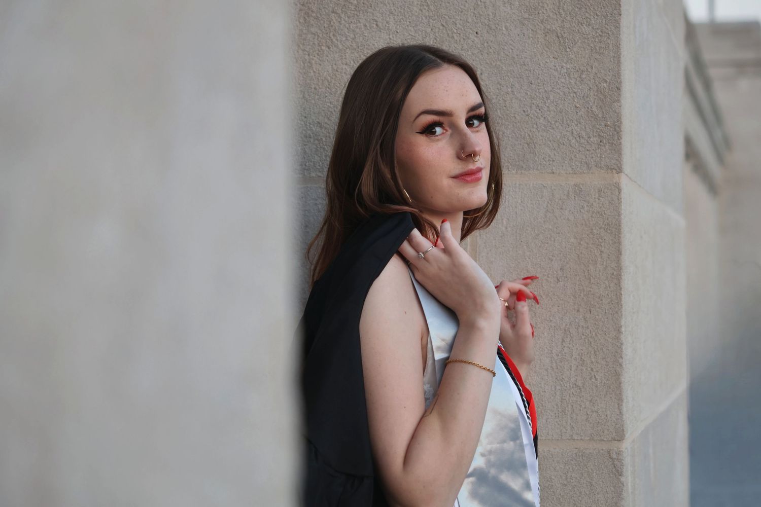 A portrait of a woman graduate posing against a stone building pillar, holding her graduation stole across her shoulders.