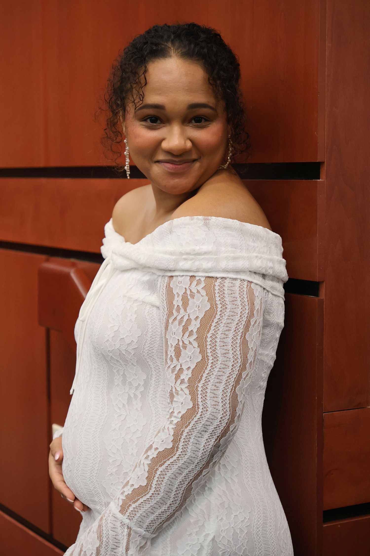 A bride smiling at the camera in an off-the-shoulder white lace dress against a warm wood-paneled wall.