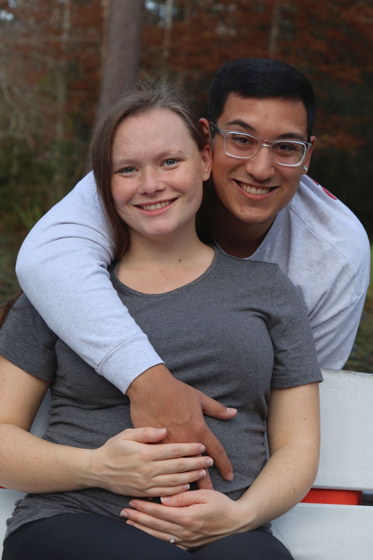 A smiling couple posing outdoors as the man wraps his arms around the woman from behind, with their hands resting over her stomach.