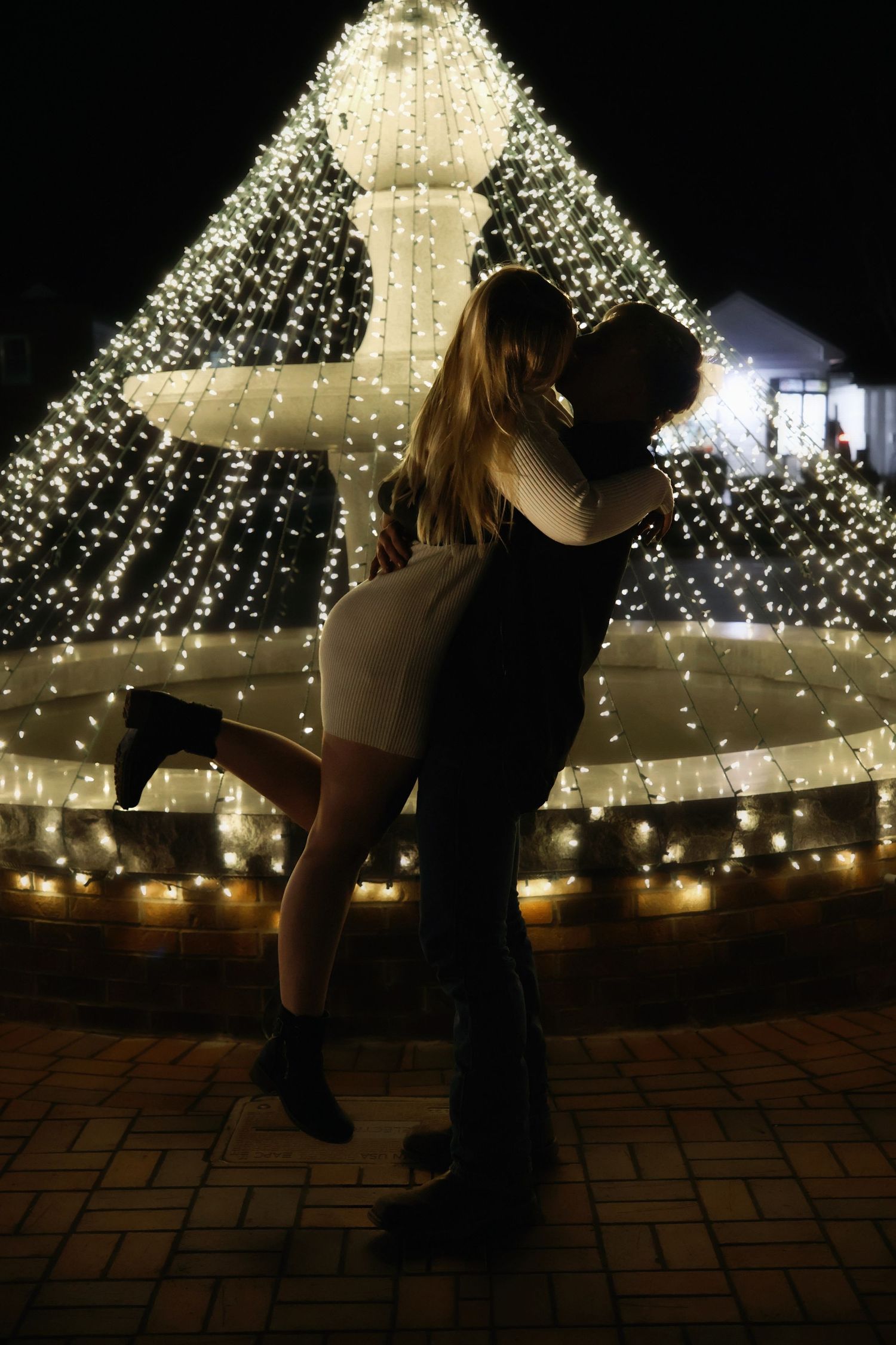 Silhouetted couple kissing in front of a brightly lit fountain at night, with the man lifting the woman as she pops one leg up.