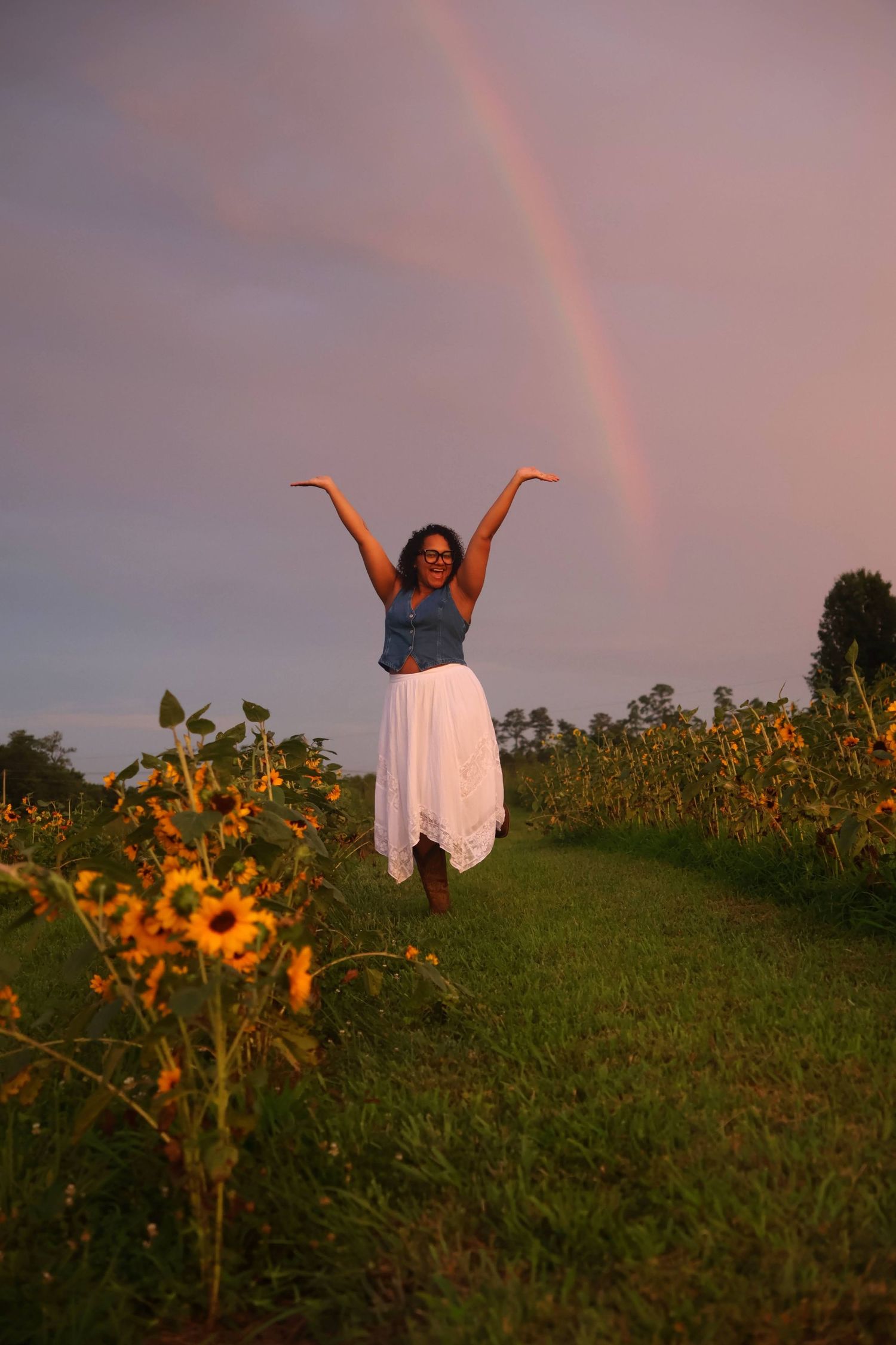 A woman  wearing glasses, a denim standing with her arms raised in a sunflower field while a rainbow is overhead.