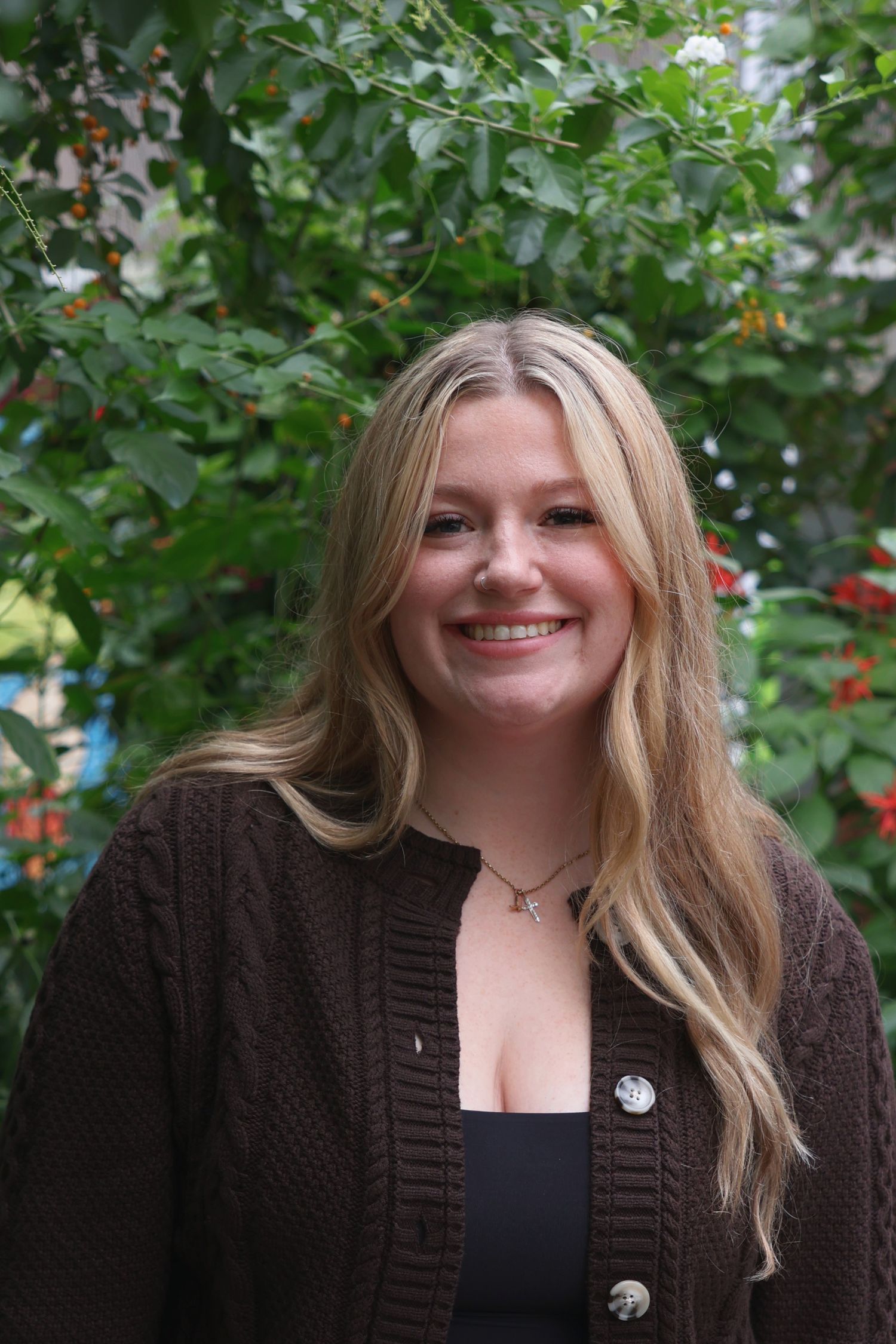 A smiling woman with blonde hair wearing a brown knit cardigan, posing outdoors in front of green foliage.