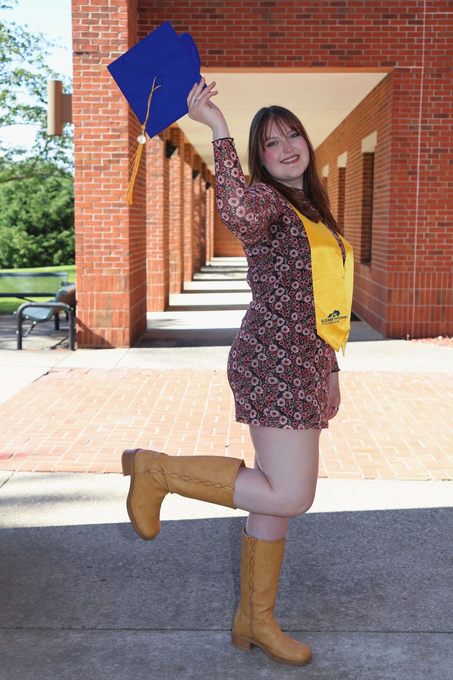 A woman holding her blue graduation cap high in the air while wearing a yellow stole, patterned dress, and tall boots near brick pillars.