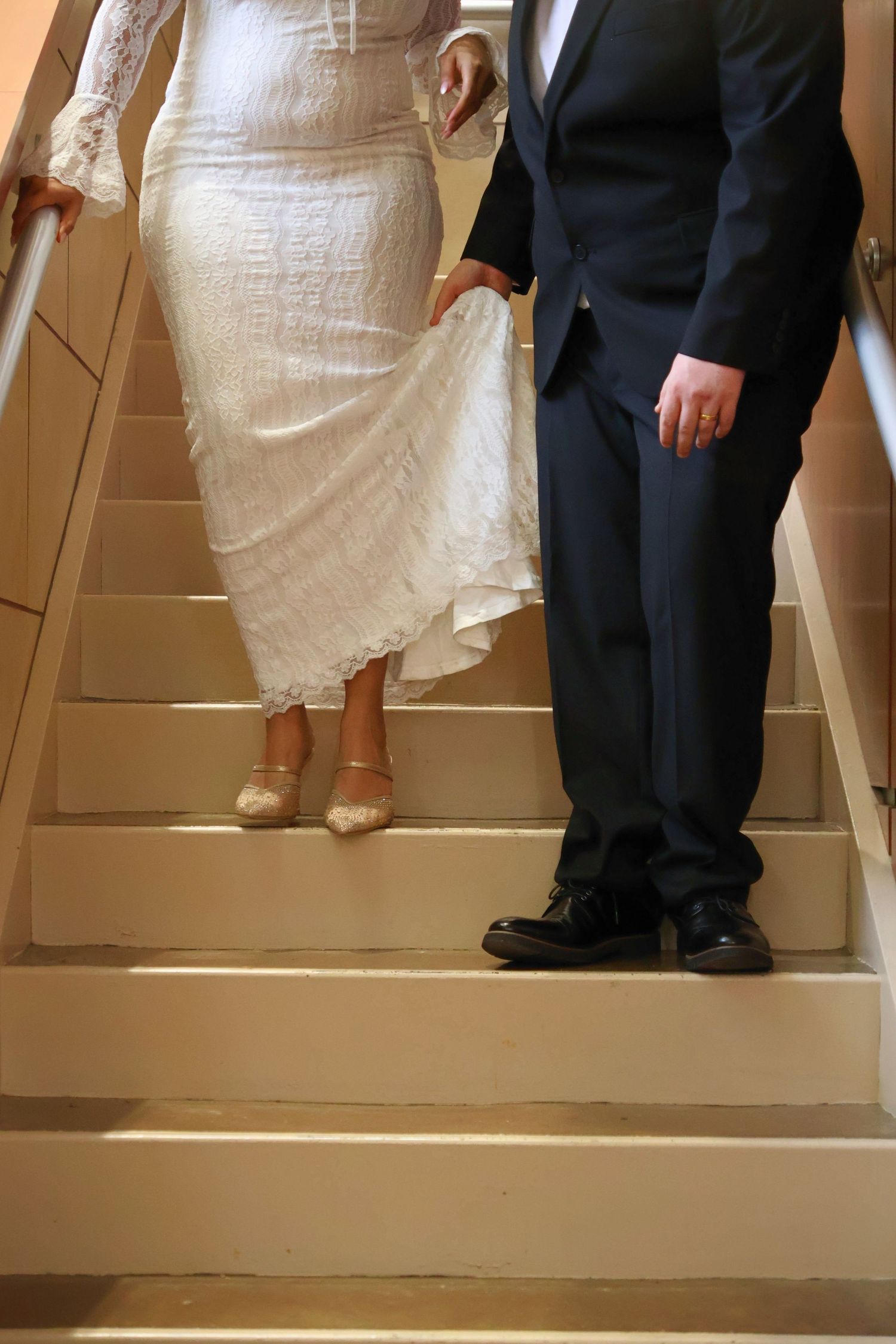 A bride in a white lace gown lifting the hem of her dress to reveal her heels while standing on a staircase next to her groom.
