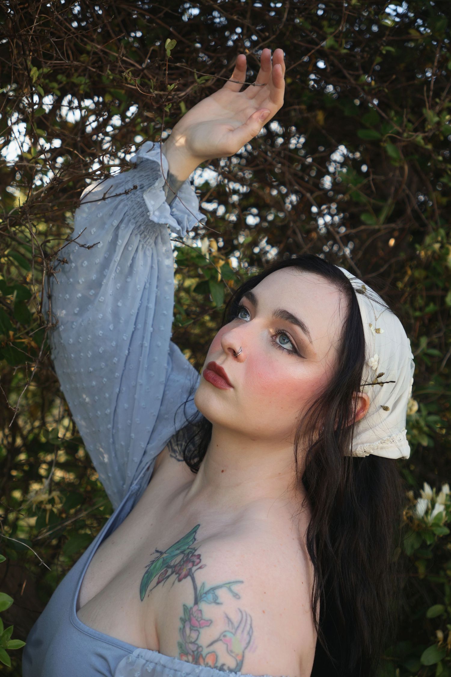 A woman wearing an embroidered bandana and off-the-shoulder blue top reaching up touching sunlight.