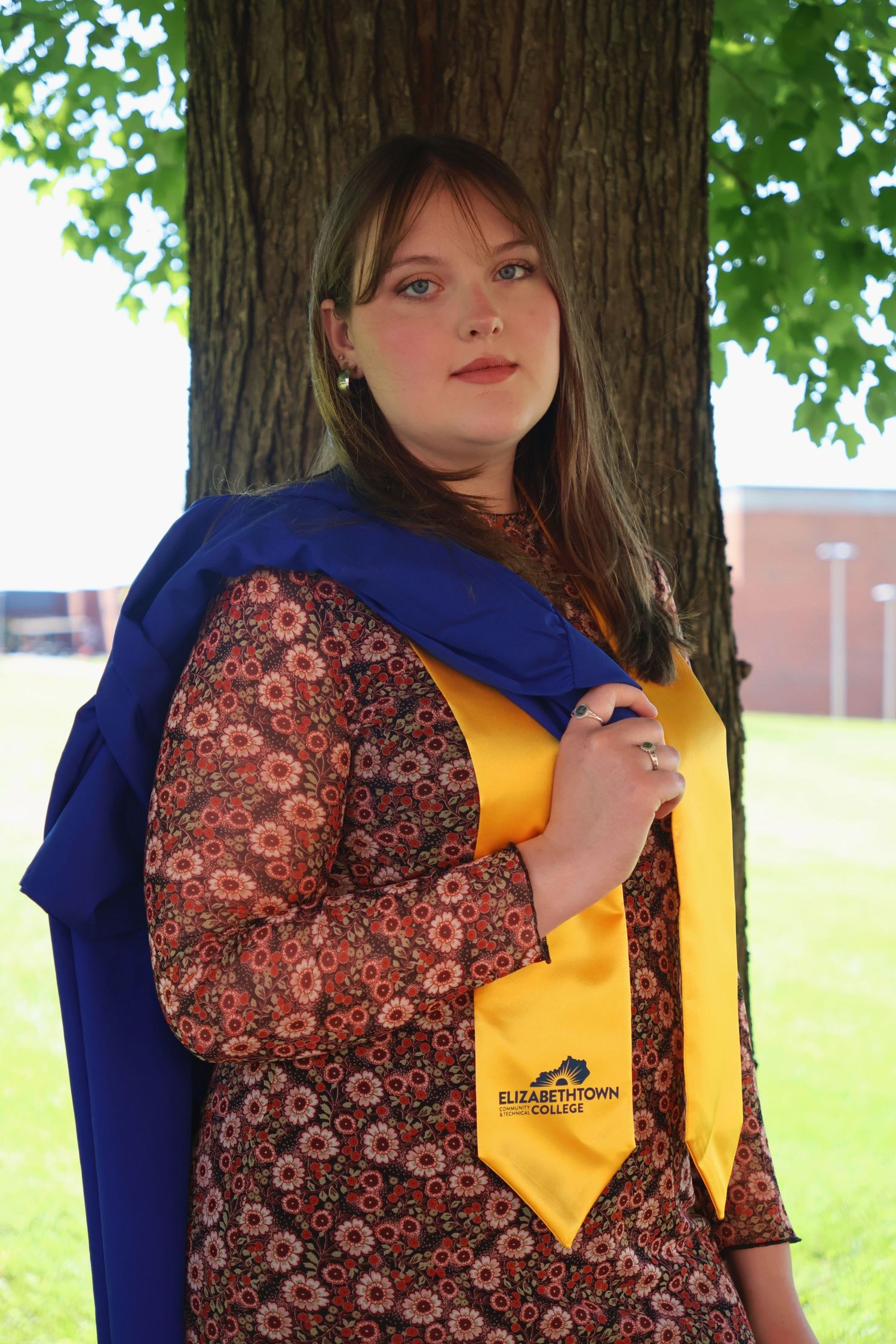 A woman leaning against a tree trunk wearing a yellow stole over a patterned dress and slinging her blue gown over her shoulder.