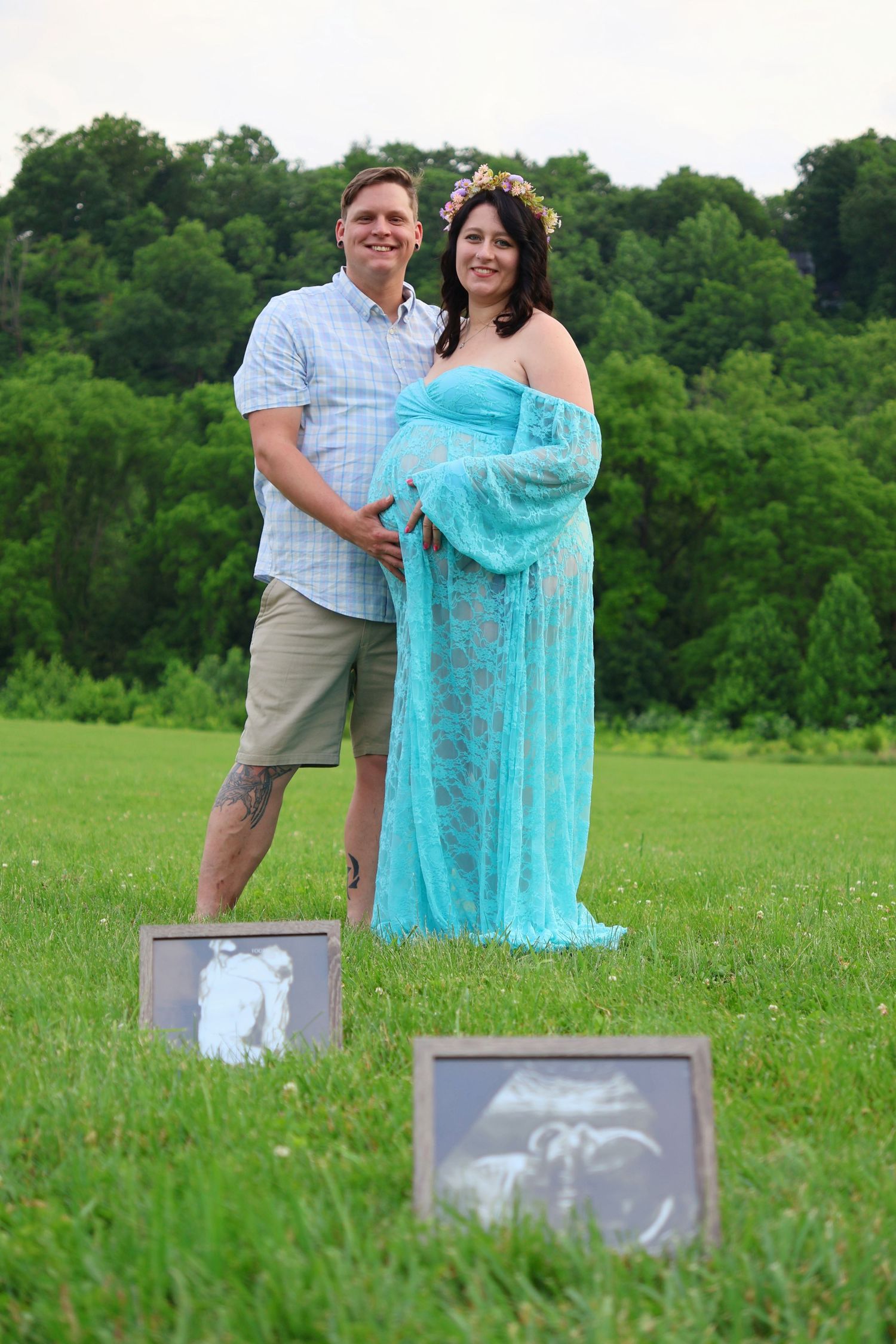 A pregnant woman in a cyan dress and a man standing together in a grassy field with two framed ultrasound photos set in front of them.