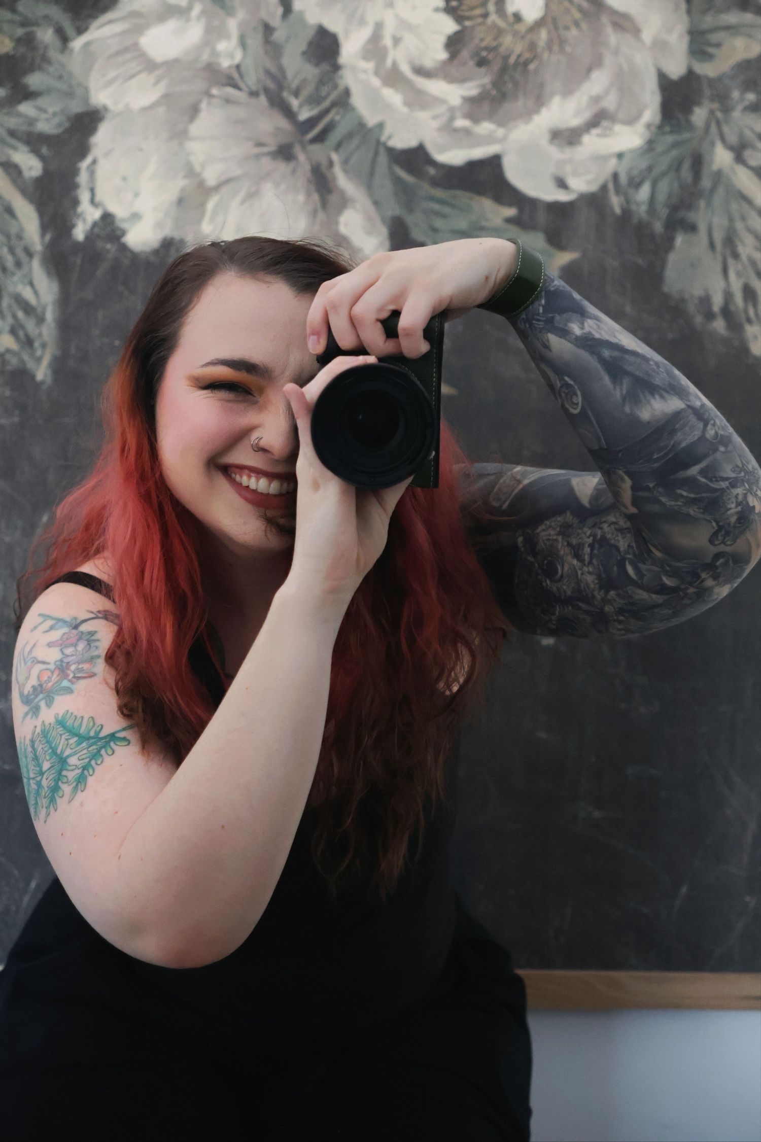 A smiling woman with dark red hair holding a camera up to her eye in a mirror reflection, set against a floral wallpaper background.
