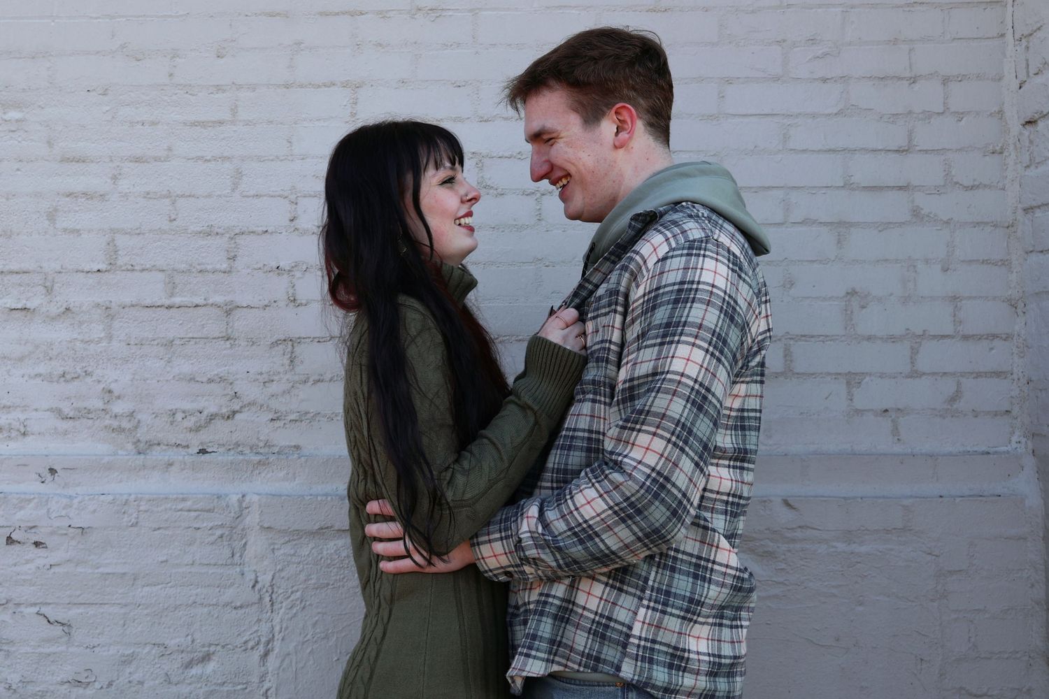 A smiling woman holding the lapels of a man's flannel shirt as they face each other against a white painted brick wall.