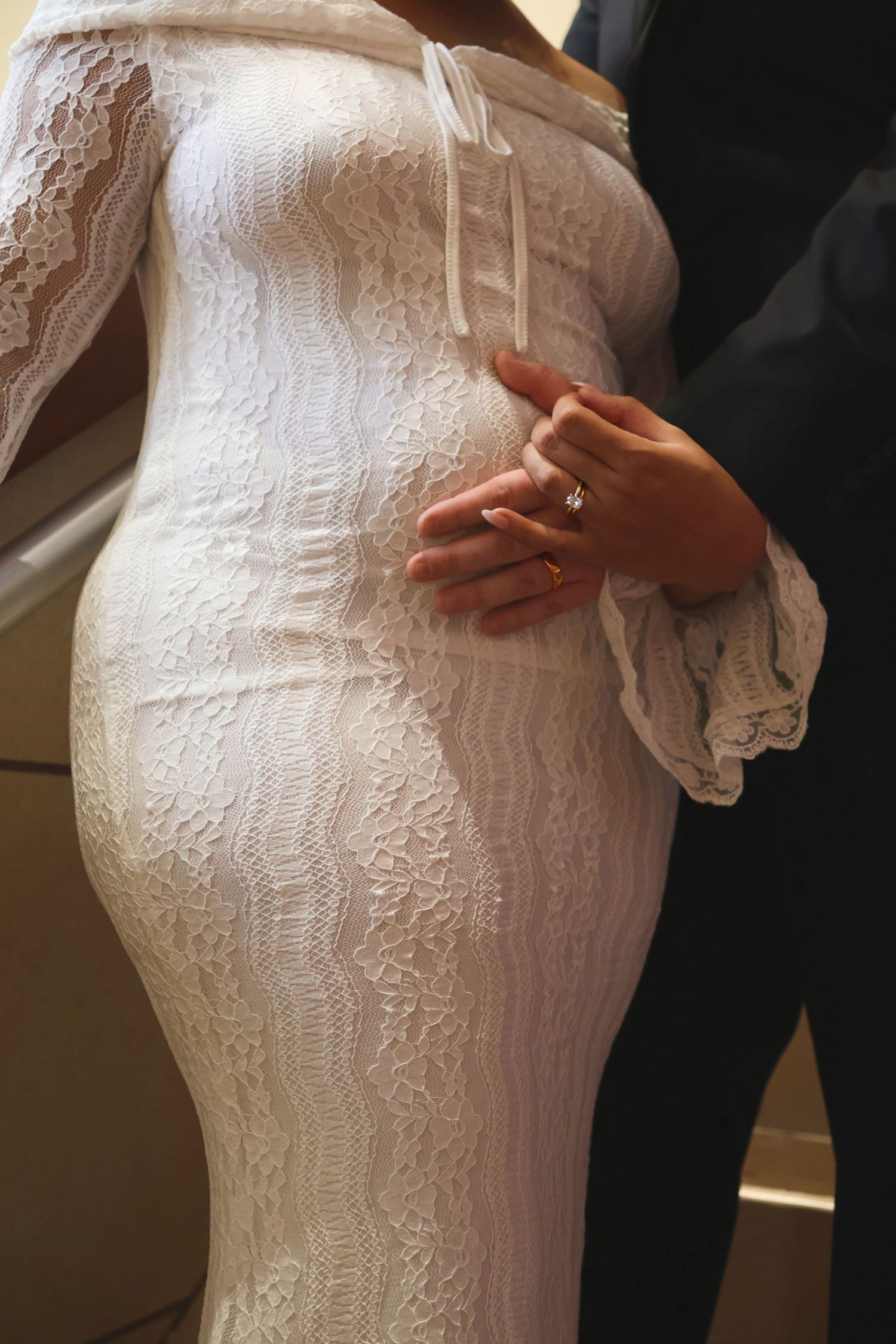 A close-up shot of a bride in a fitted white lace dress with her hand gently resting over her stomach.