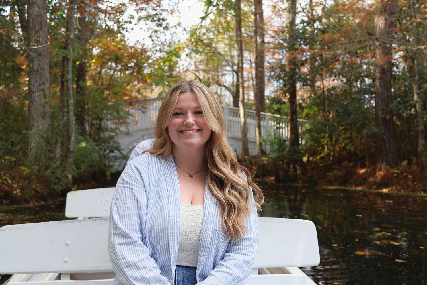 A smiling woman with blonde hair sitting on a a boat with autumn trees in the background.