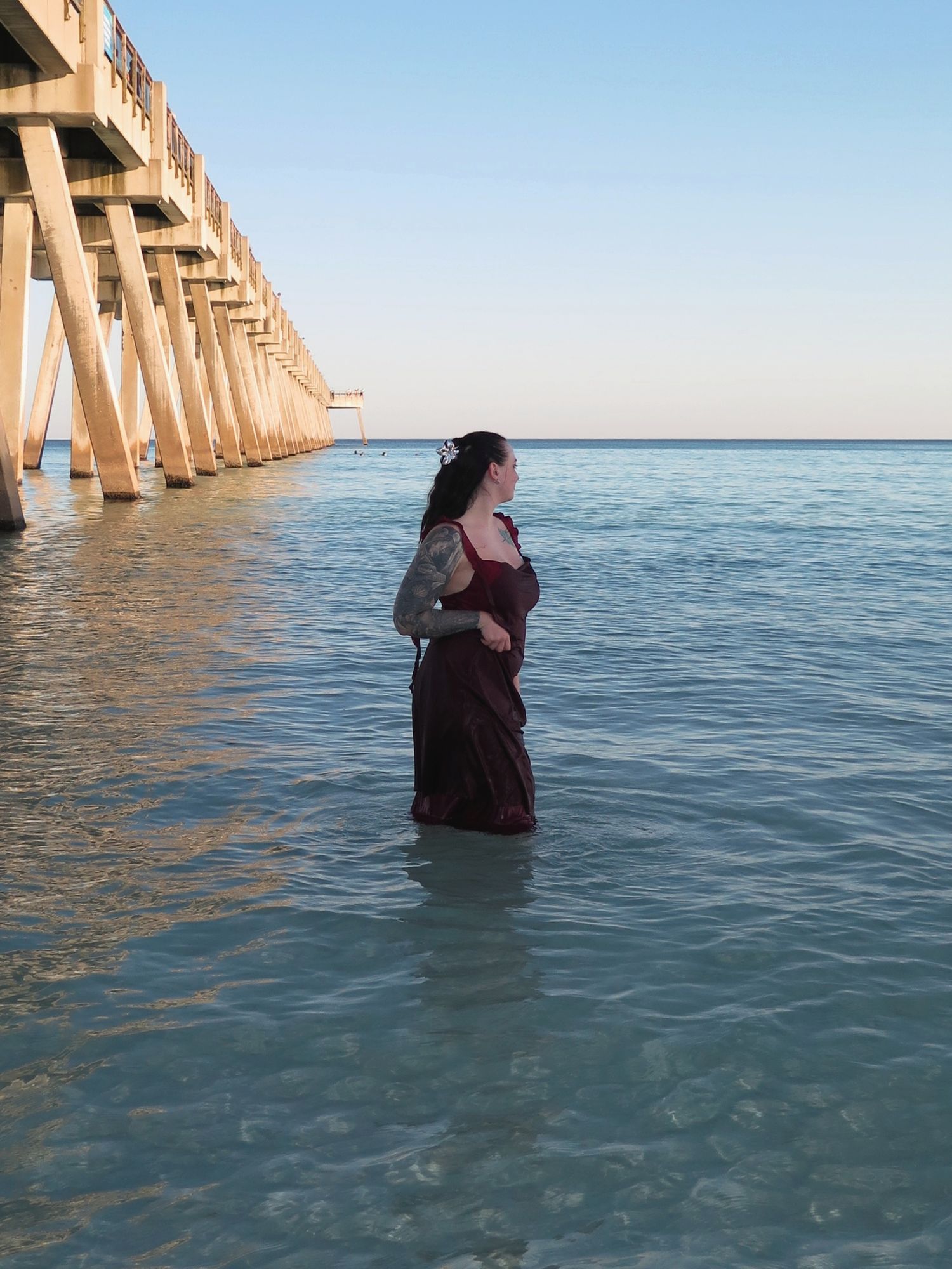 A woman in a maroon dress facing the horizon while standing waist-deep in the ocean next to a wooden pier.