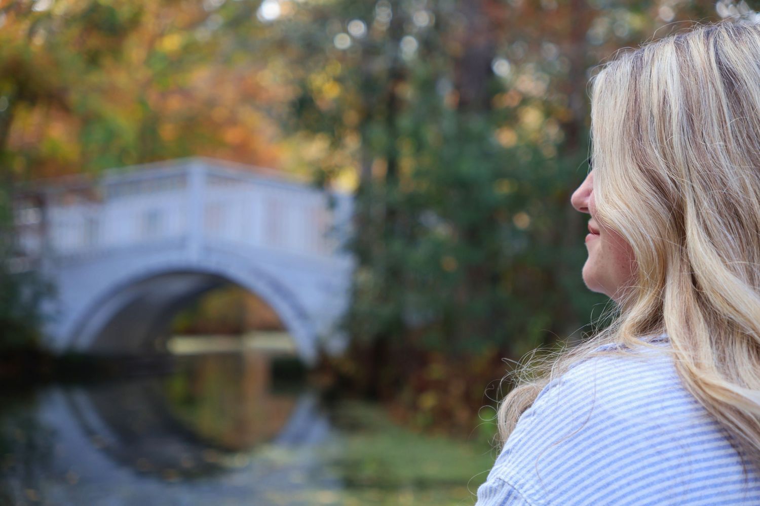 Side profile of a blonde woman in a boat looking toward an arched footbridge over water surrounded by autumn foliage.
