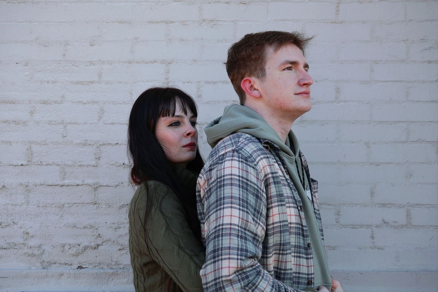 A young woman with dark hair leaning gently against a man in a plaid flannel shirt, both looking off into the distance against a white paint