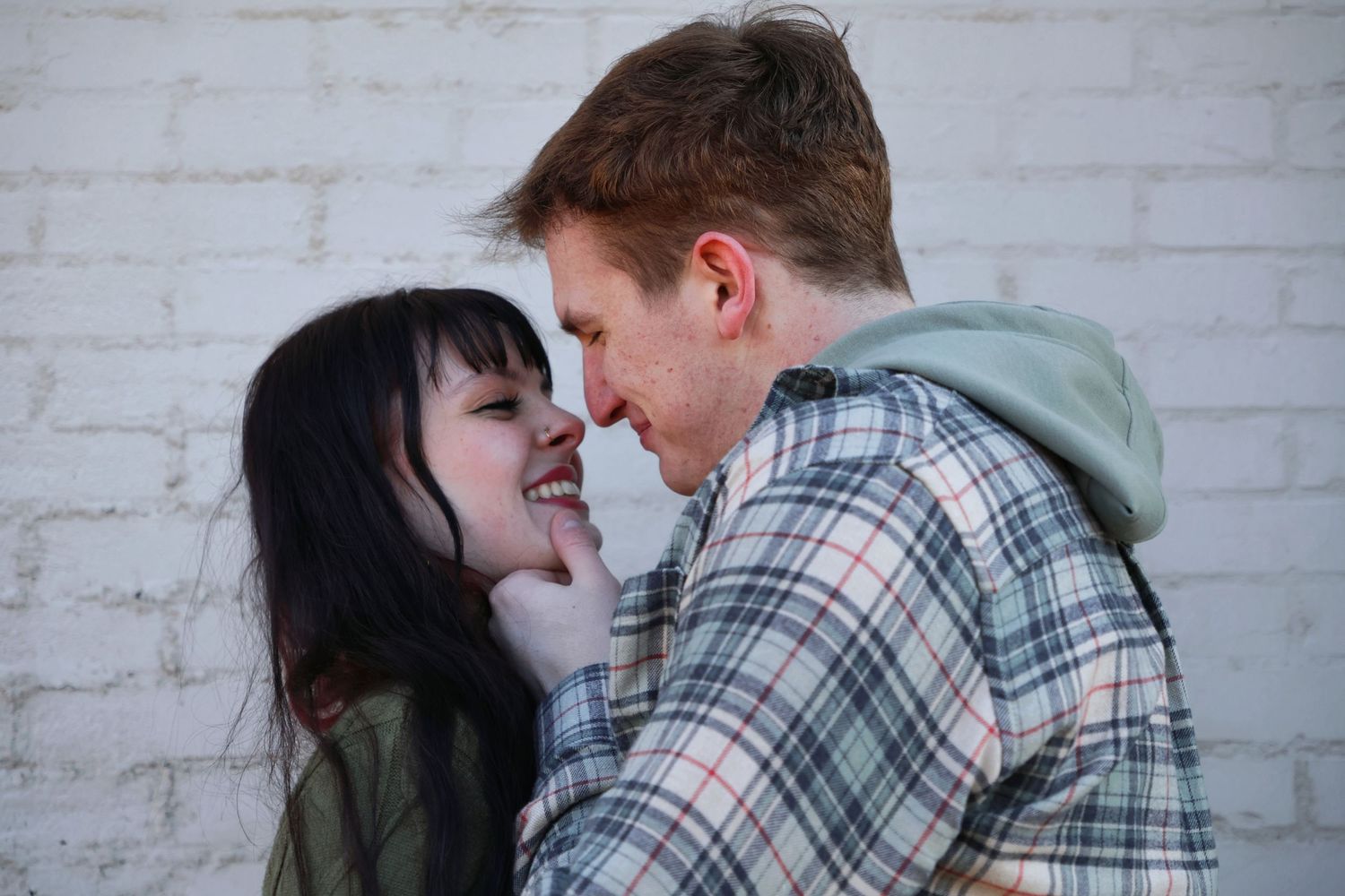 A close-up portrait of a man gently holding a woman's chin as they look into each other's eyes against a white painted brick wall.