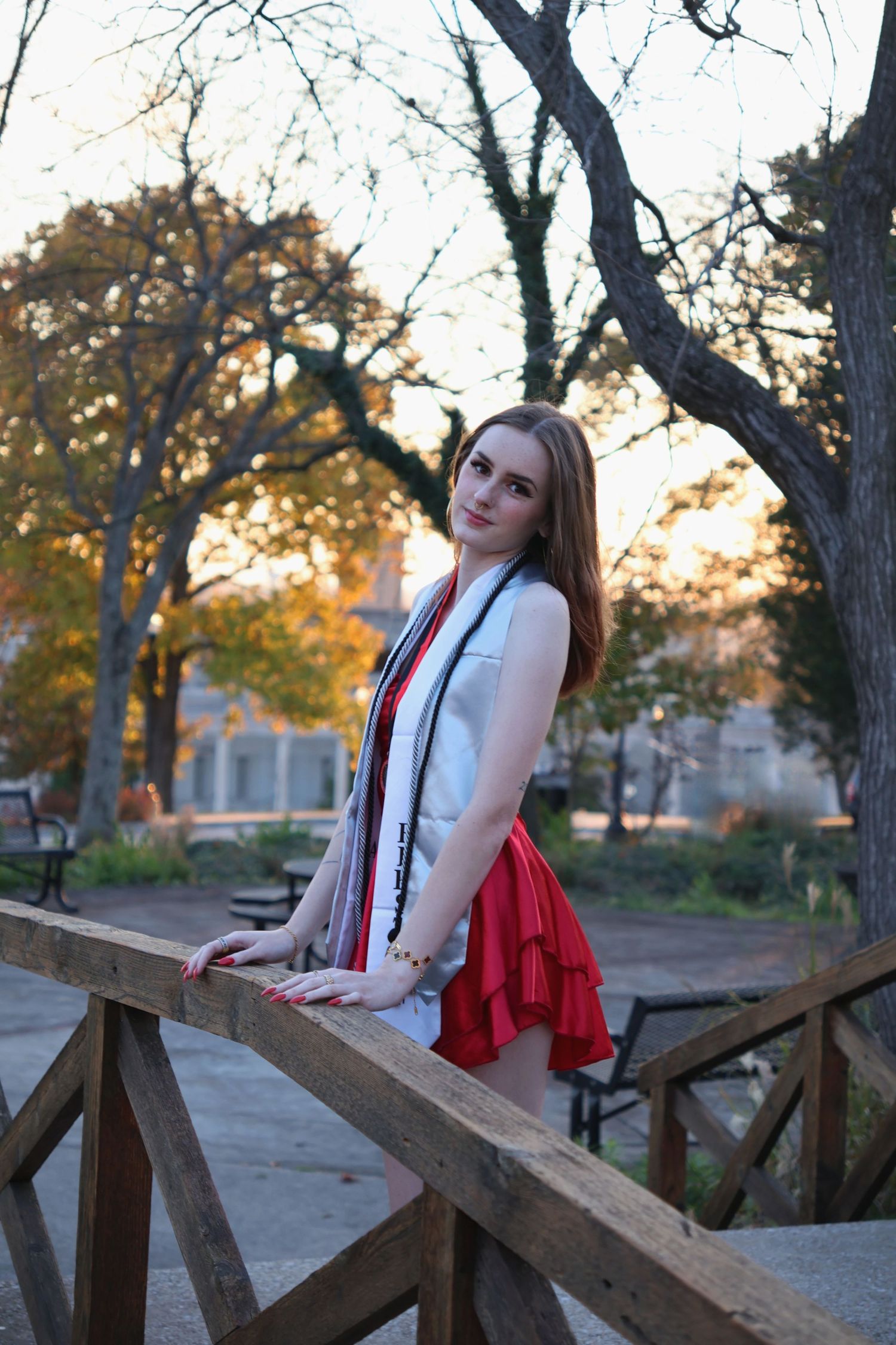 A graduating woman wearing a stole leaning against a wooden bridge railing with autumn trees in the background.