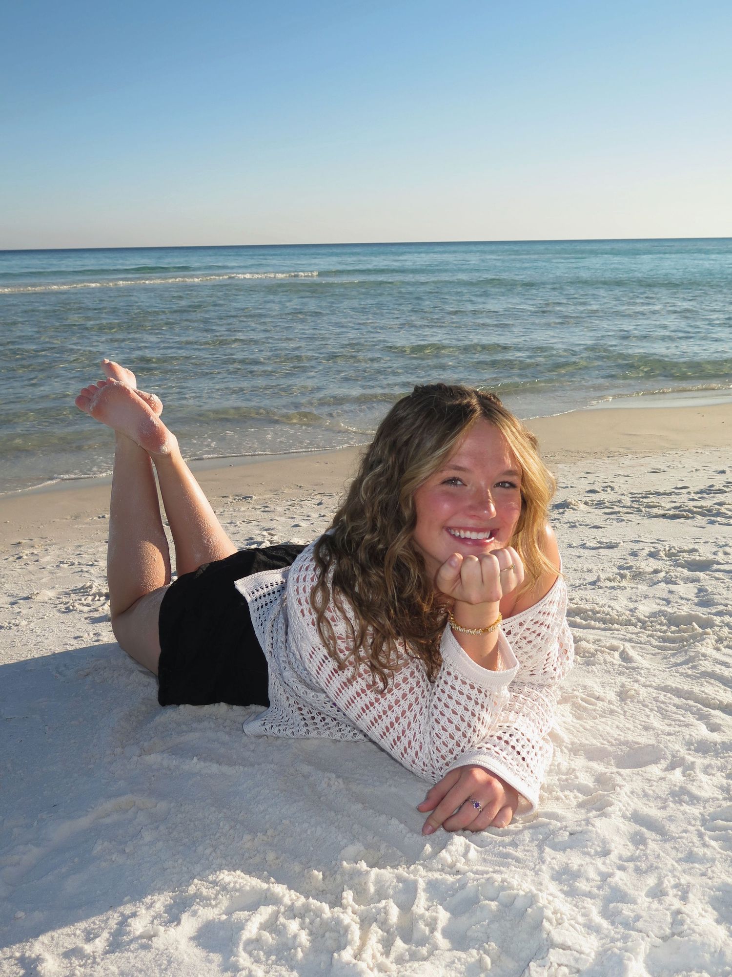 A smiling woman lying on her stomach in white sand along the beach, resting her chin on her hands.