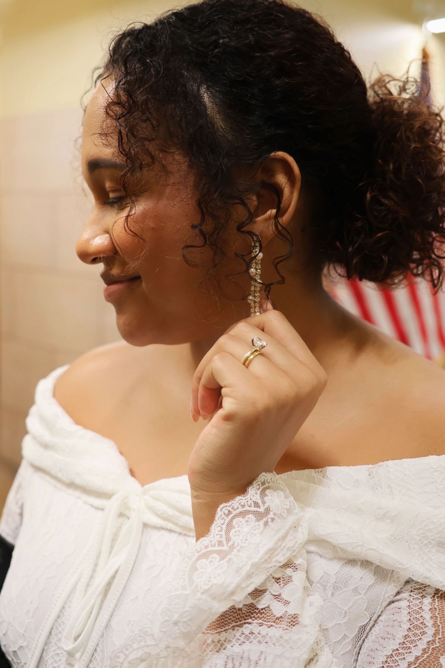 Side profile of a bride in an off-the-shoulder lace dress gently touching her drop earring with her hand raised.