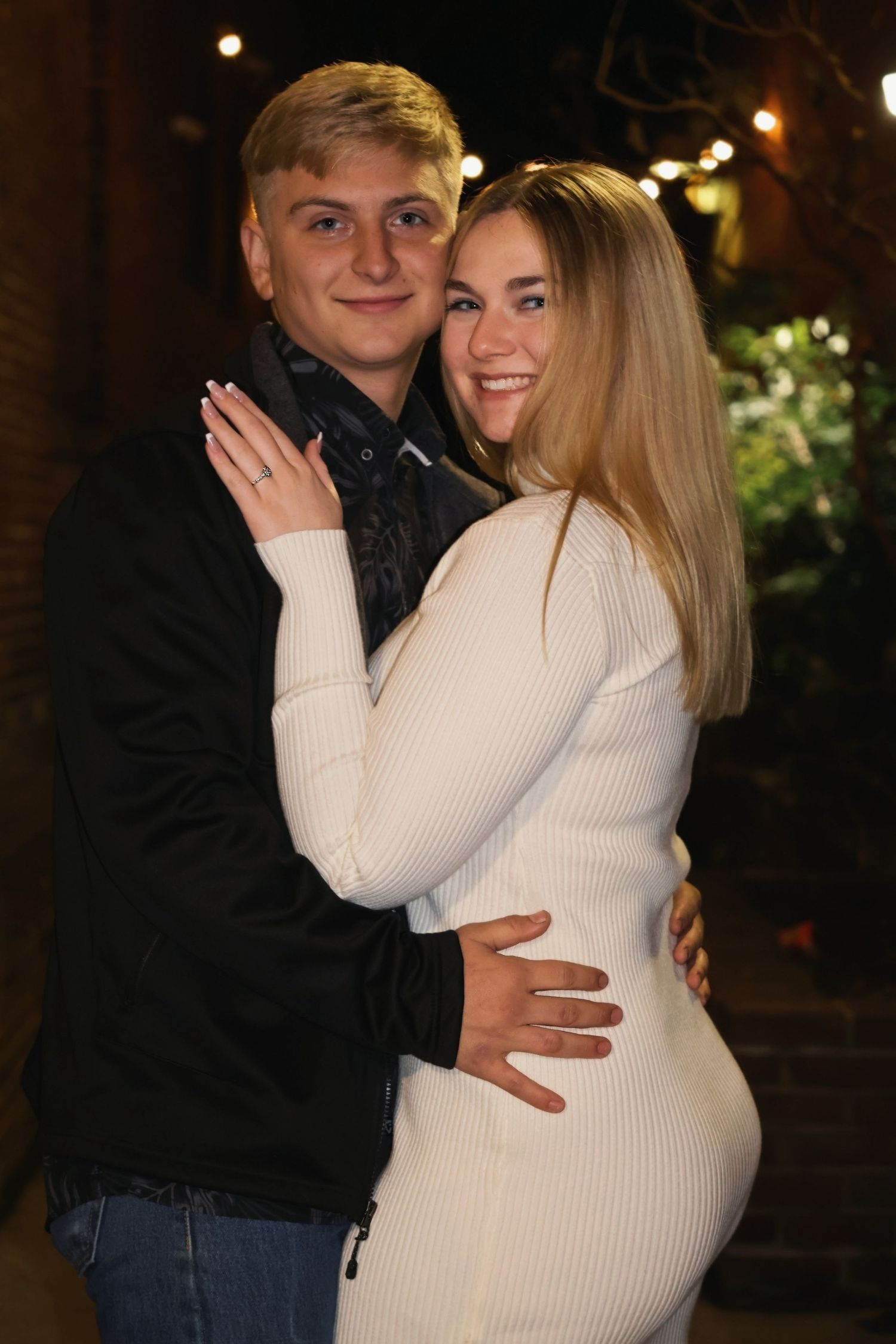 A smiling young couple embracing outdoors at night, with the woman in a white long-sleeve dress and the man in a dark jacket.