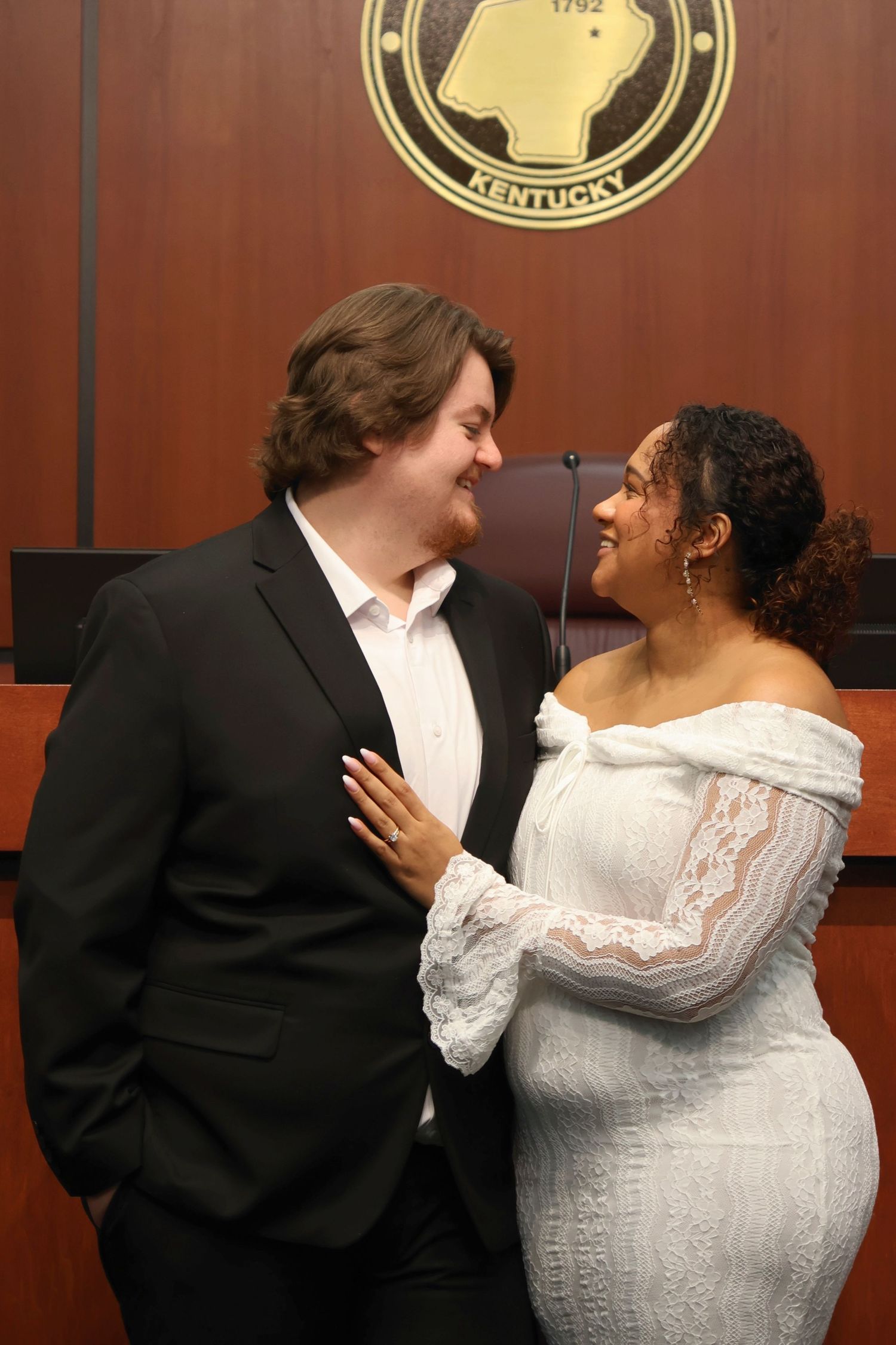 A bride in a lace off-the-shoulder dress and a groom in a dark suit embracing and looking at each other during a courtroom ceremony.