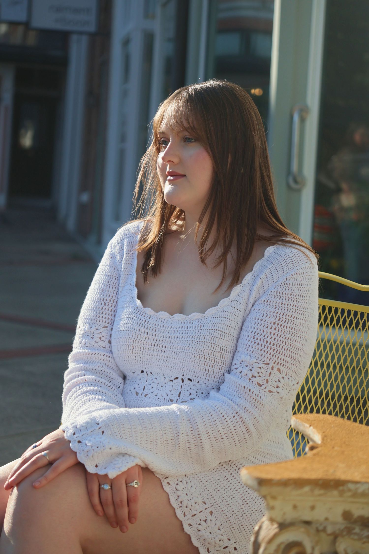 A woman in a white long-sleeve knit dress sitting outdoors on a patio chair, looking off to the side.