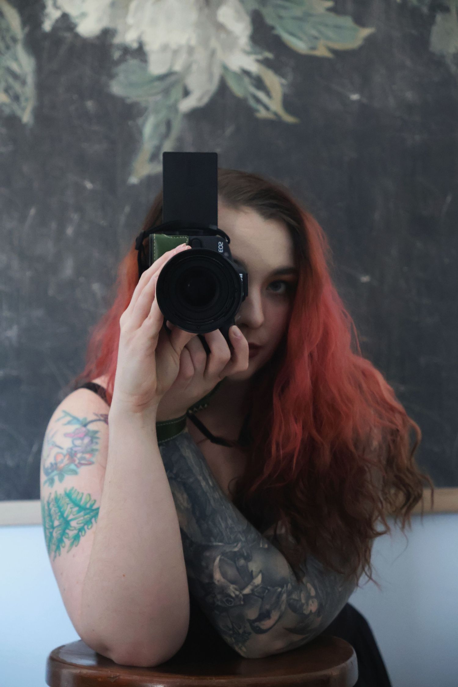 A woman with dark and red hair taking a mirror selfie while holding a camera up to cover part of her face.