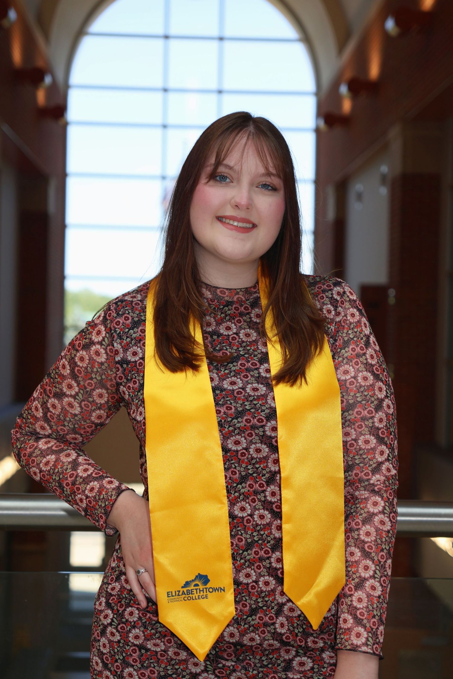 A portrait of a smiling woman in a yellow graduation stole and patterned dress posing indoors in front of large windows.