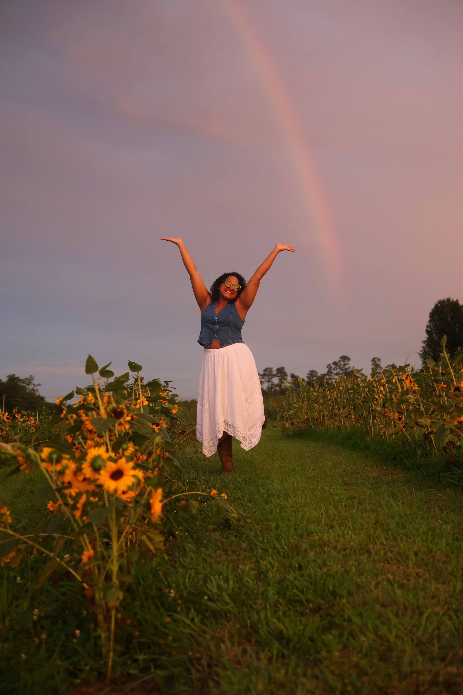 A woman with glasses & a denim top standing in a sunflower field with her arms raised in joy beneath a rainbow stretching across a dusk sky.