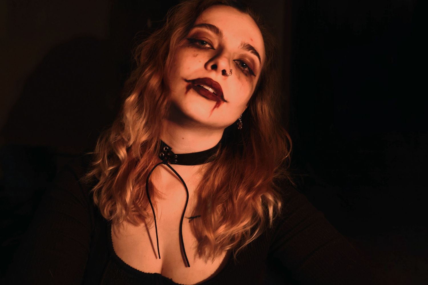 In low light, a woman wearing a black choker tilts her head with dark eye makeup and smudged red lipstick trailing down her chin.