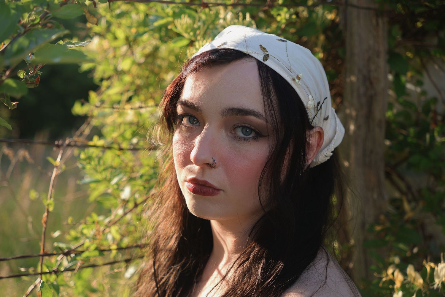 A woman wearing an embroidered bandana sits during golden hour with a barbed wire fence behind her.
