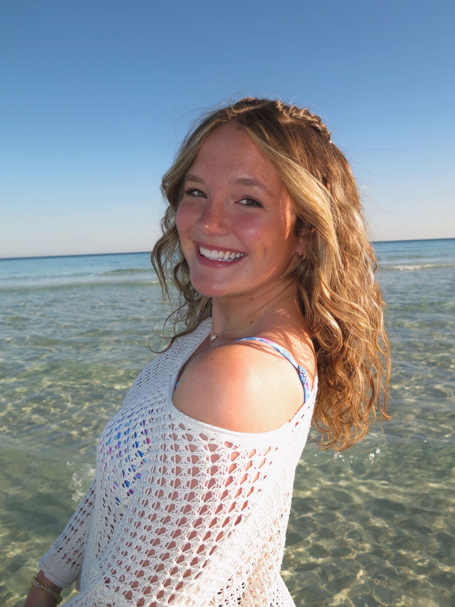 A portrait of a smiling woman with wavy blonde hair on a beach with clear water behind her during sunny weather.
