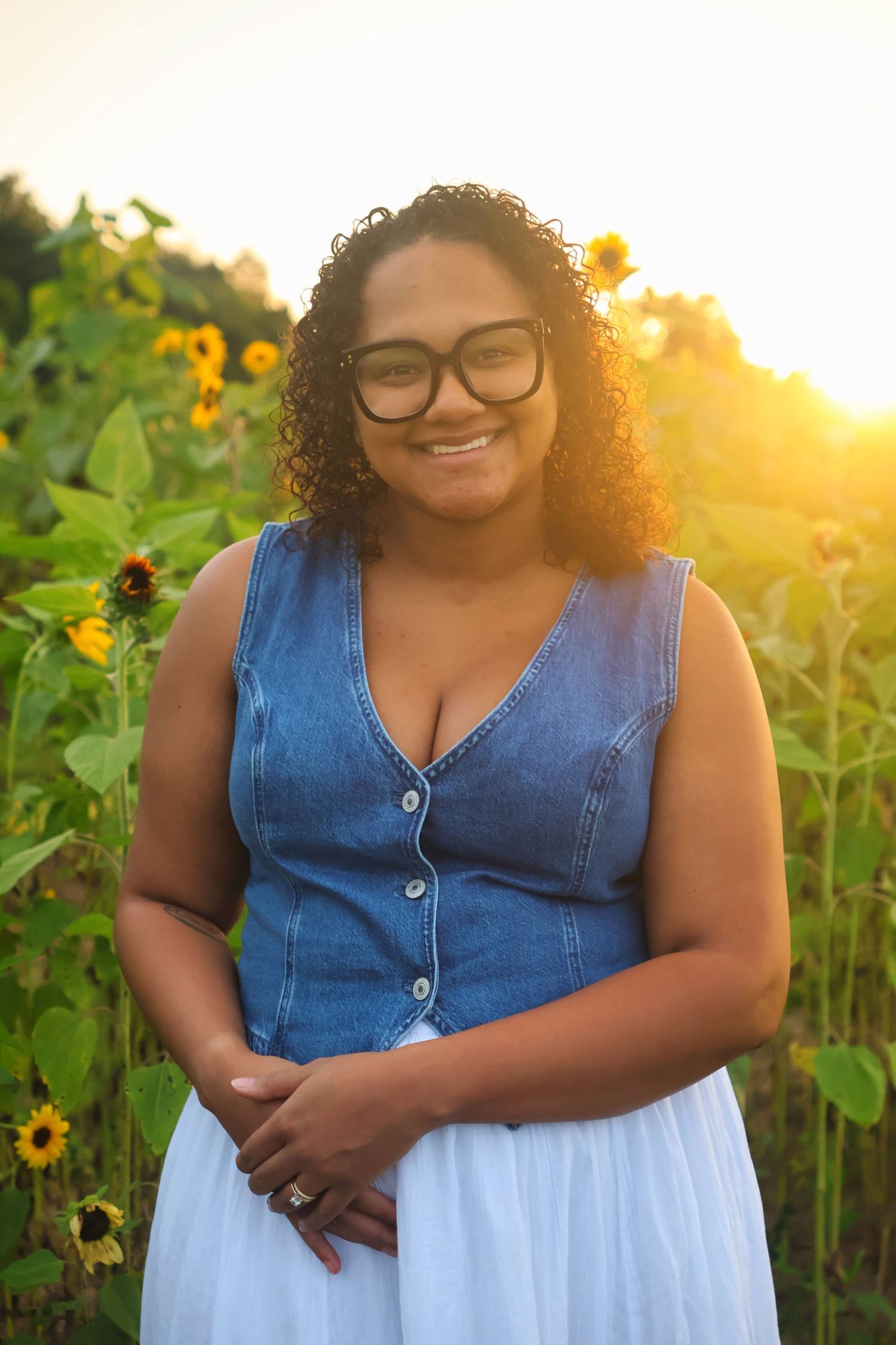 A smiling woman wearing glasses, a denim top, and a white skirt standing in a sunflower field during golden hour backlight.