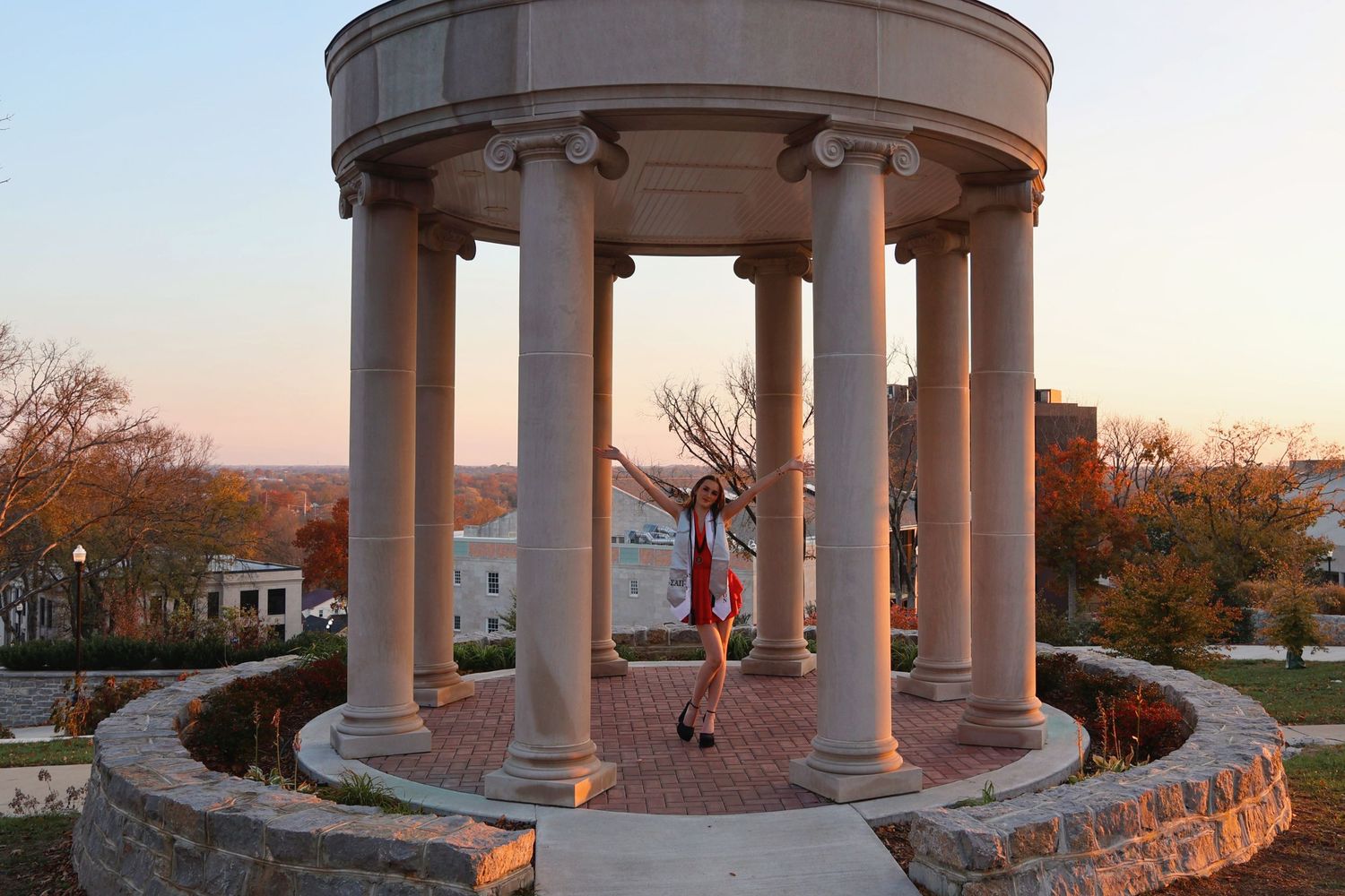 A wide shot of a woman posing inside a large stone rotunda during sunset with autumnal trees in the background wearing graduation regalia.