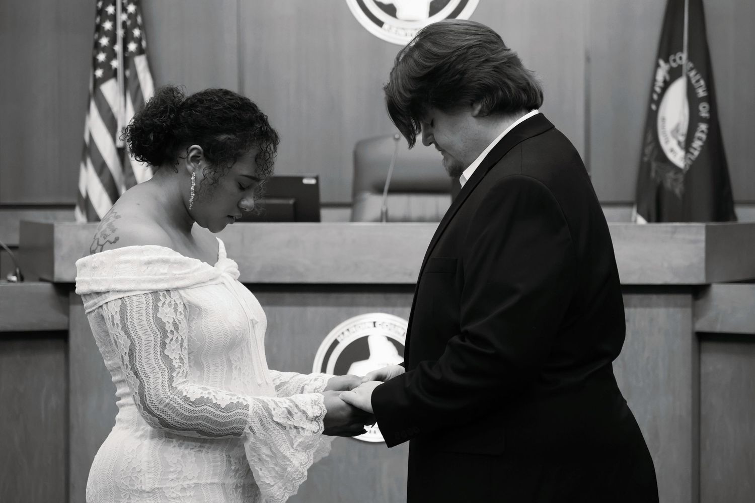 Black and white photograph of a bride and groom holding hands with their heads bowed down during a prayer in a courtroom ceremony.