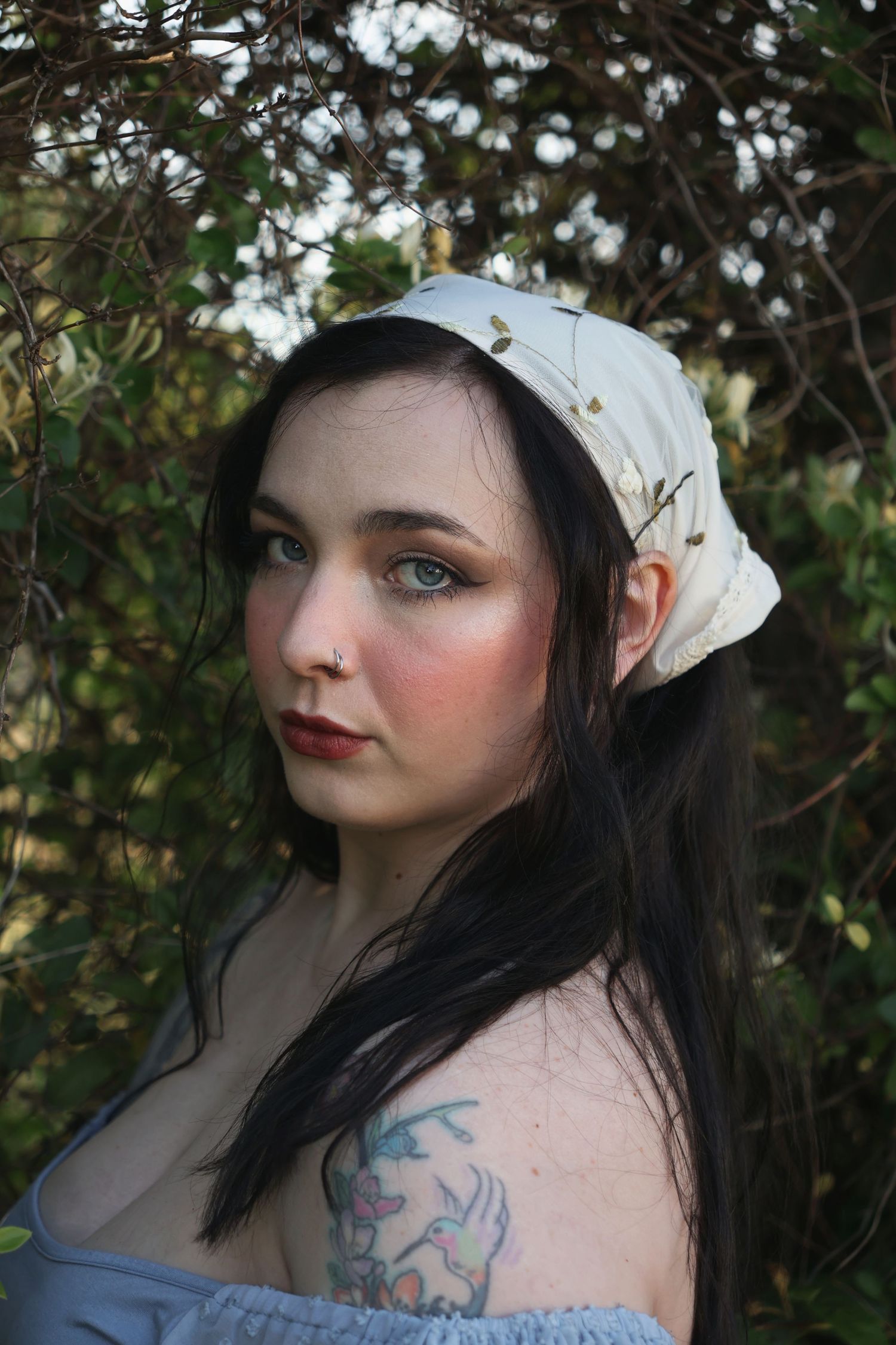A woman wearing an embroidered bandana and off-the-shoulder blue top making eye contact with the camera.