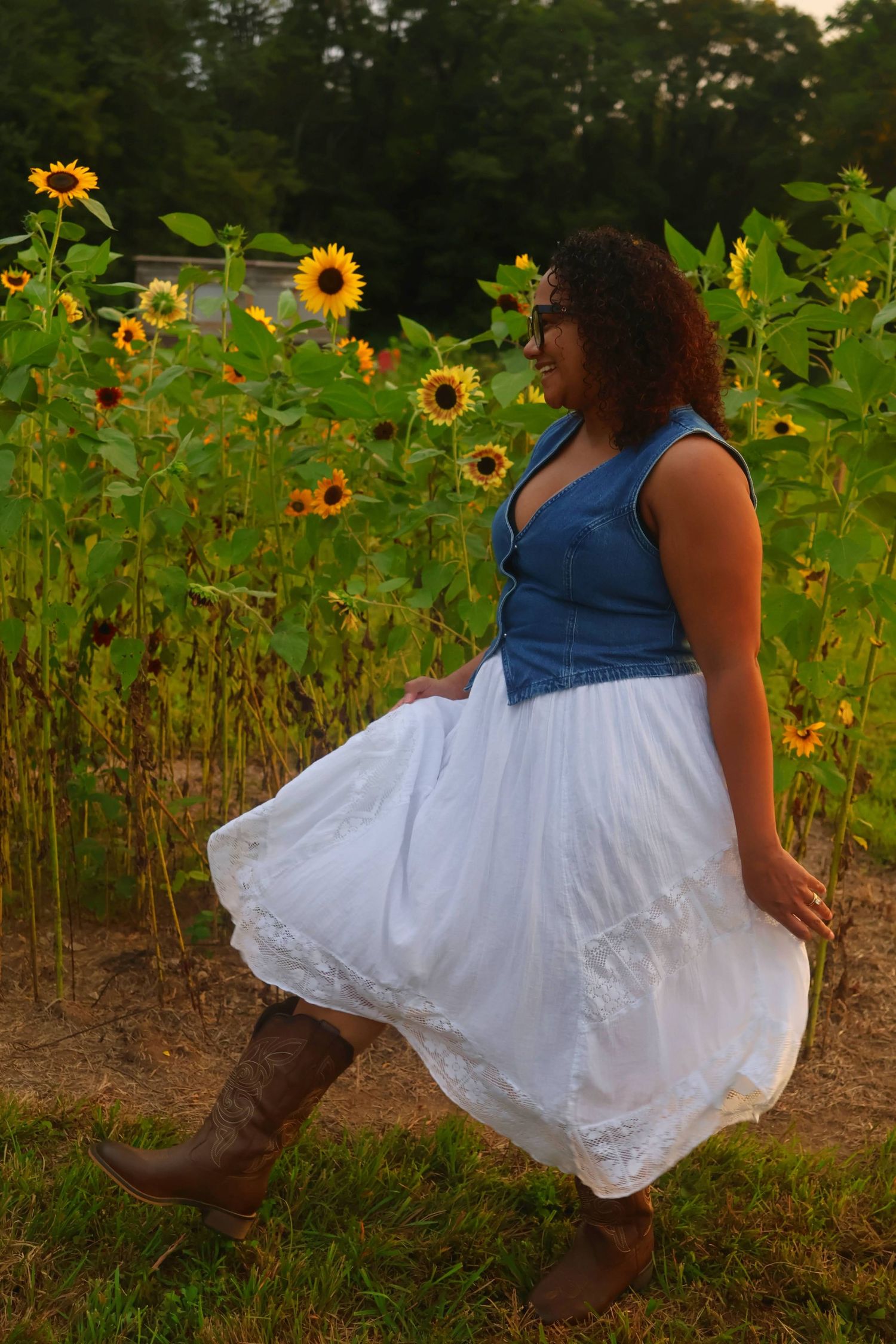 A woman  wearing glasses, and a denim top running through a sunflower field with a swishing white skirt and boots.